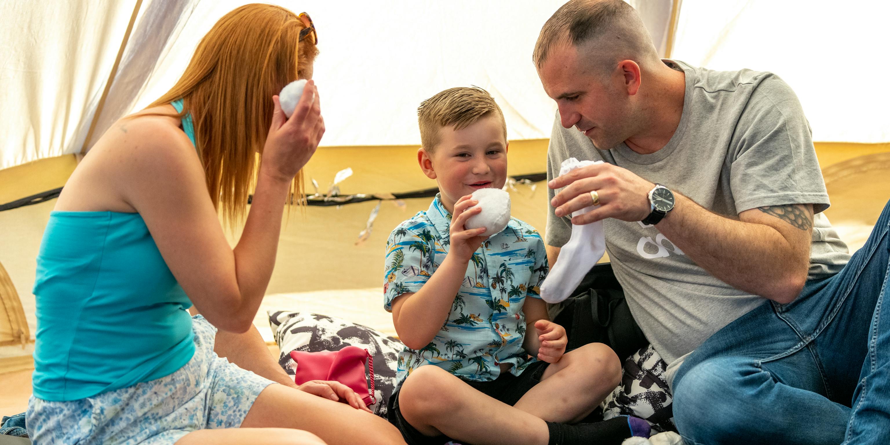 A family sat together on the floor. The parent/carer to the left is looking at the young child in the middle with a white ball in their hand.  The other parent/carer is looking at the child with a sock in their hand. The young child in the middle is smiling and holding a white sock/ball