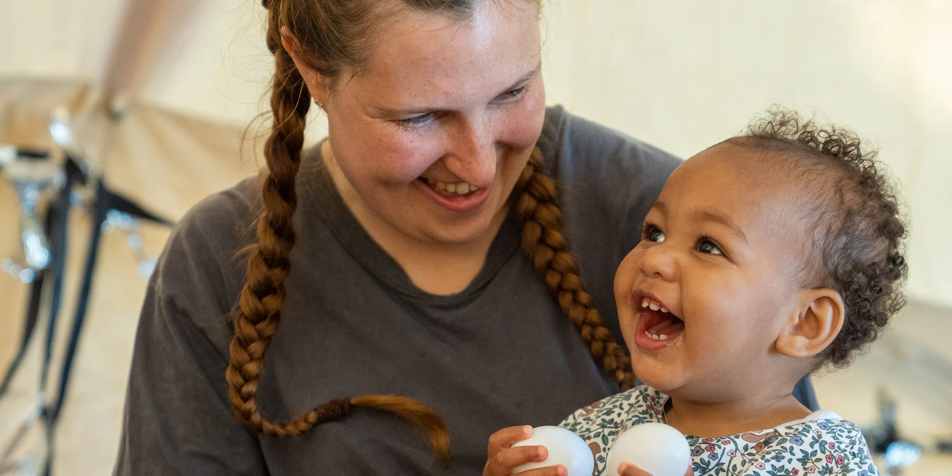 A young baby with their parent/carer are sat together. The baby is holding two white balls in their hands, their parent/carer is holding up the other. They are both smiling and laughing.