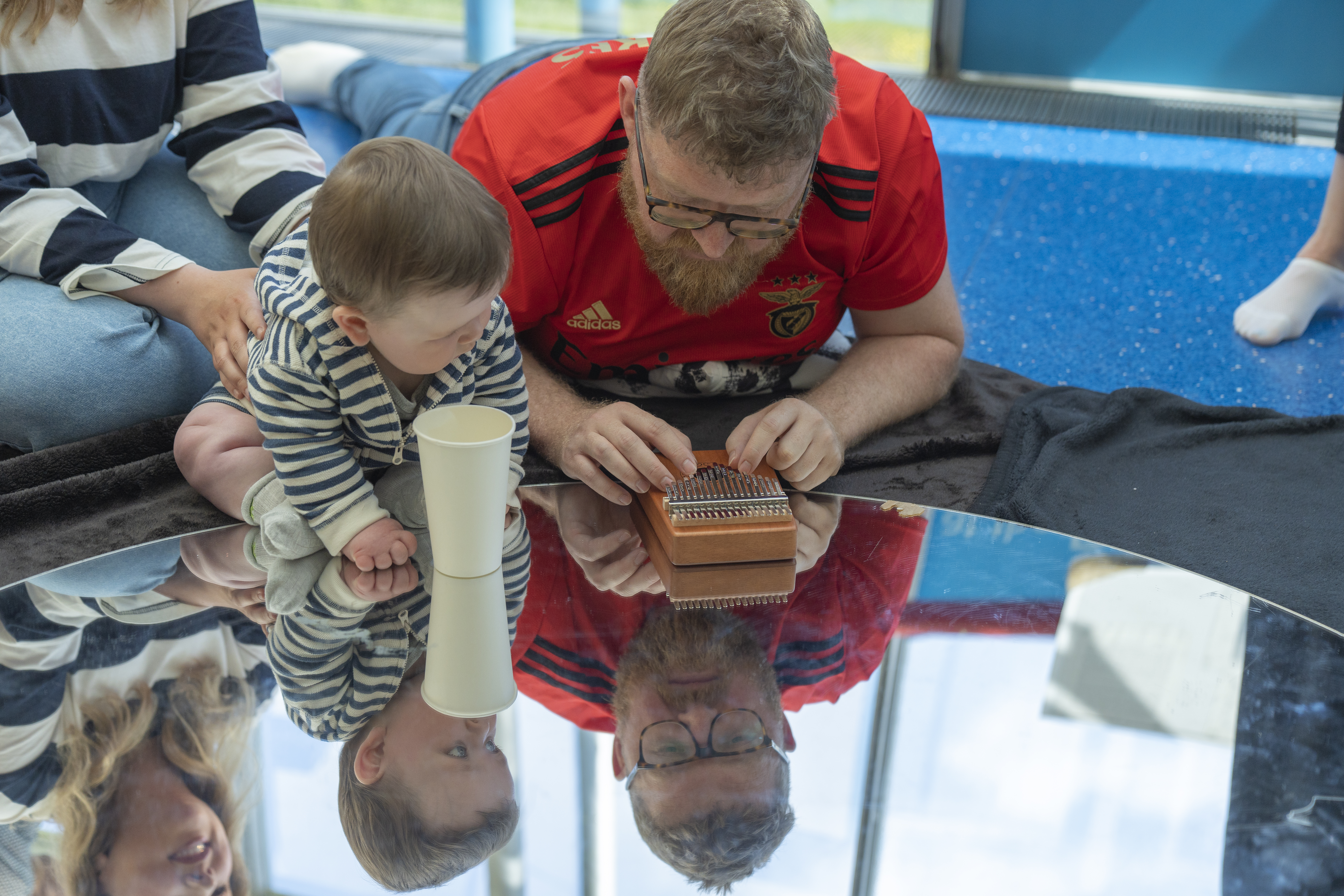 A baby is sat on top of a mirror. Next to them is their parent/carer who is playing with a musical instrument. The baby is looking at their parent/carer playing with this instrument 