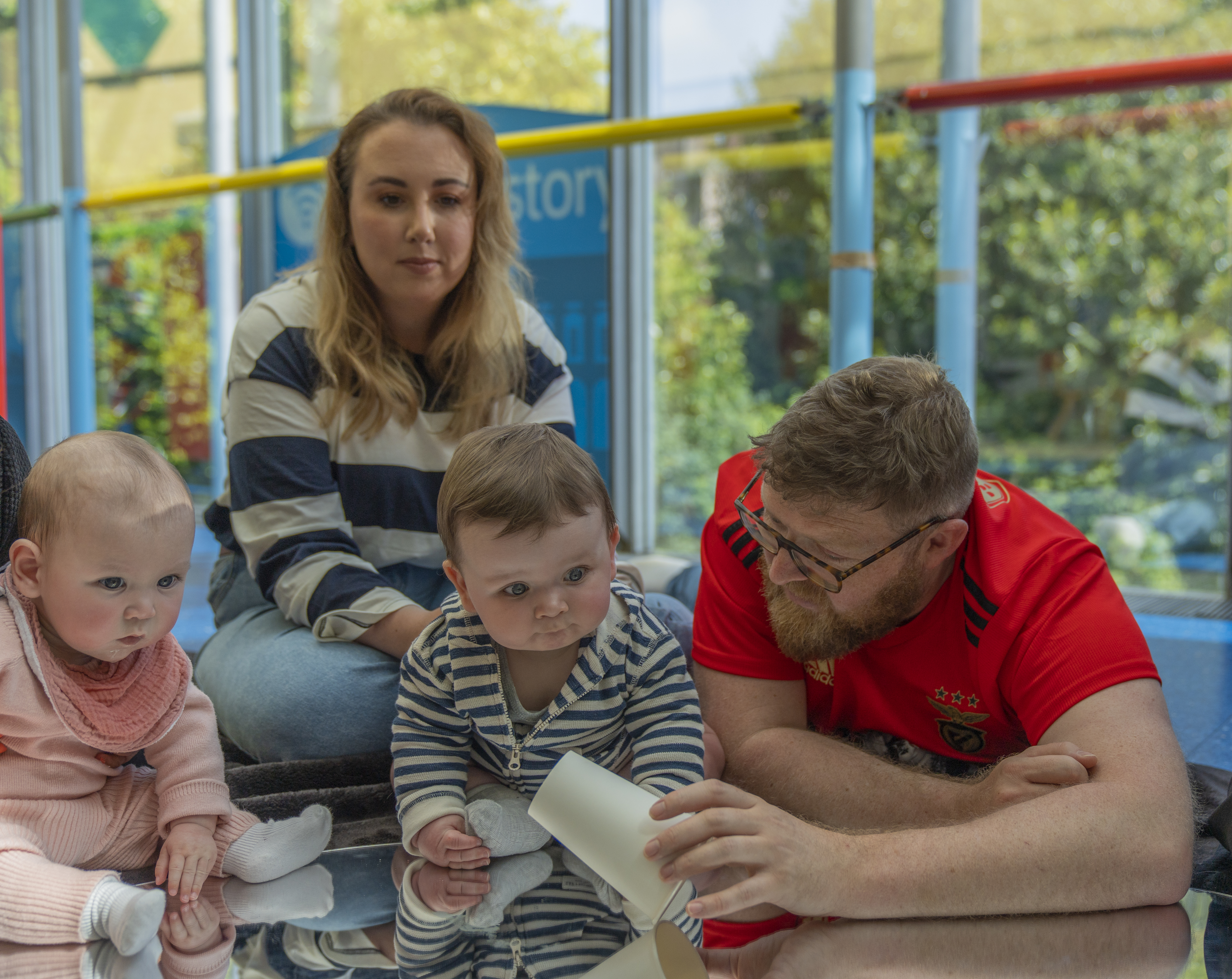 A baby is sat on top of a black mat and mirror. Next to them is their parent/carer who is playing with a musical instrument. The baby is looking at their parent/carer playing with this instrument 