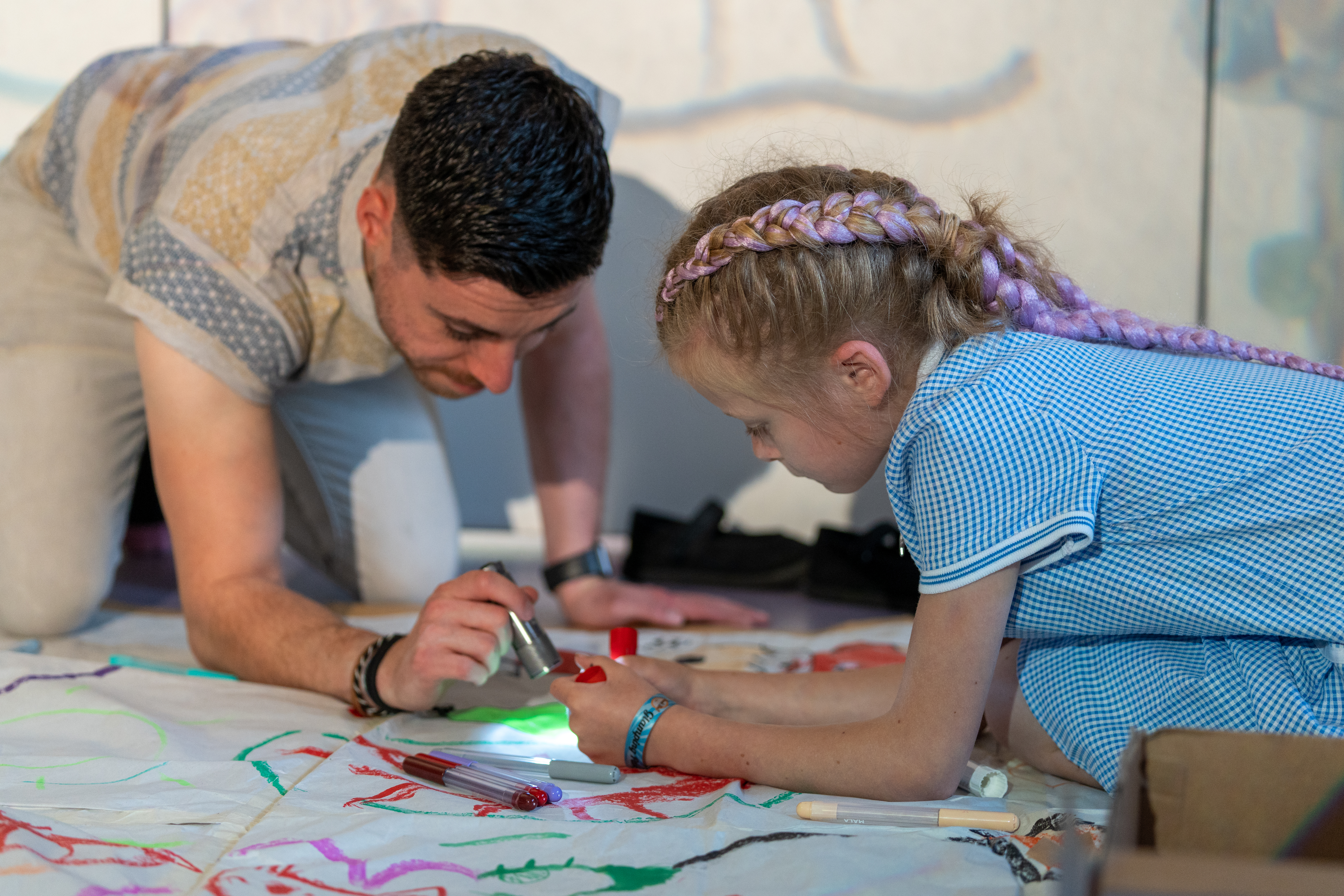 An artist is kneeling on the floor next to a young child. The artist is shining a torch at a something the young child is holding. 