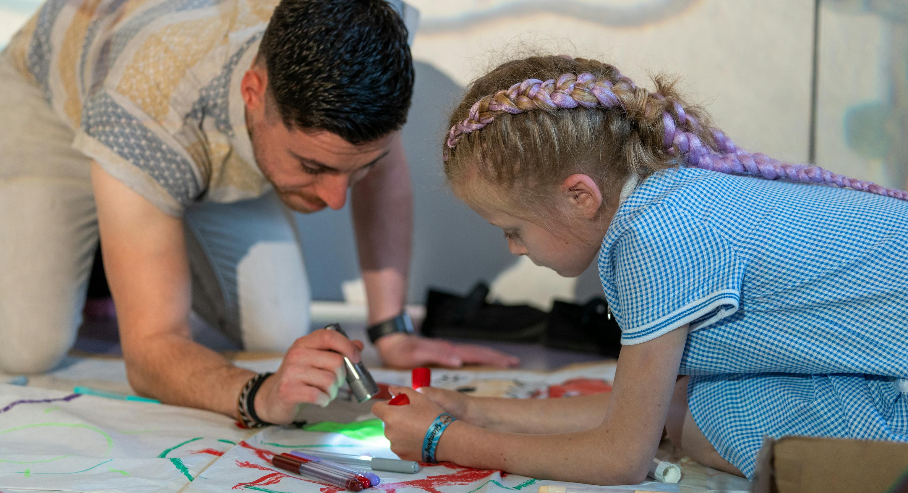 An artist is kneeling on the floor next to a young child. The artist is shining a torch at a something the young child is holding.