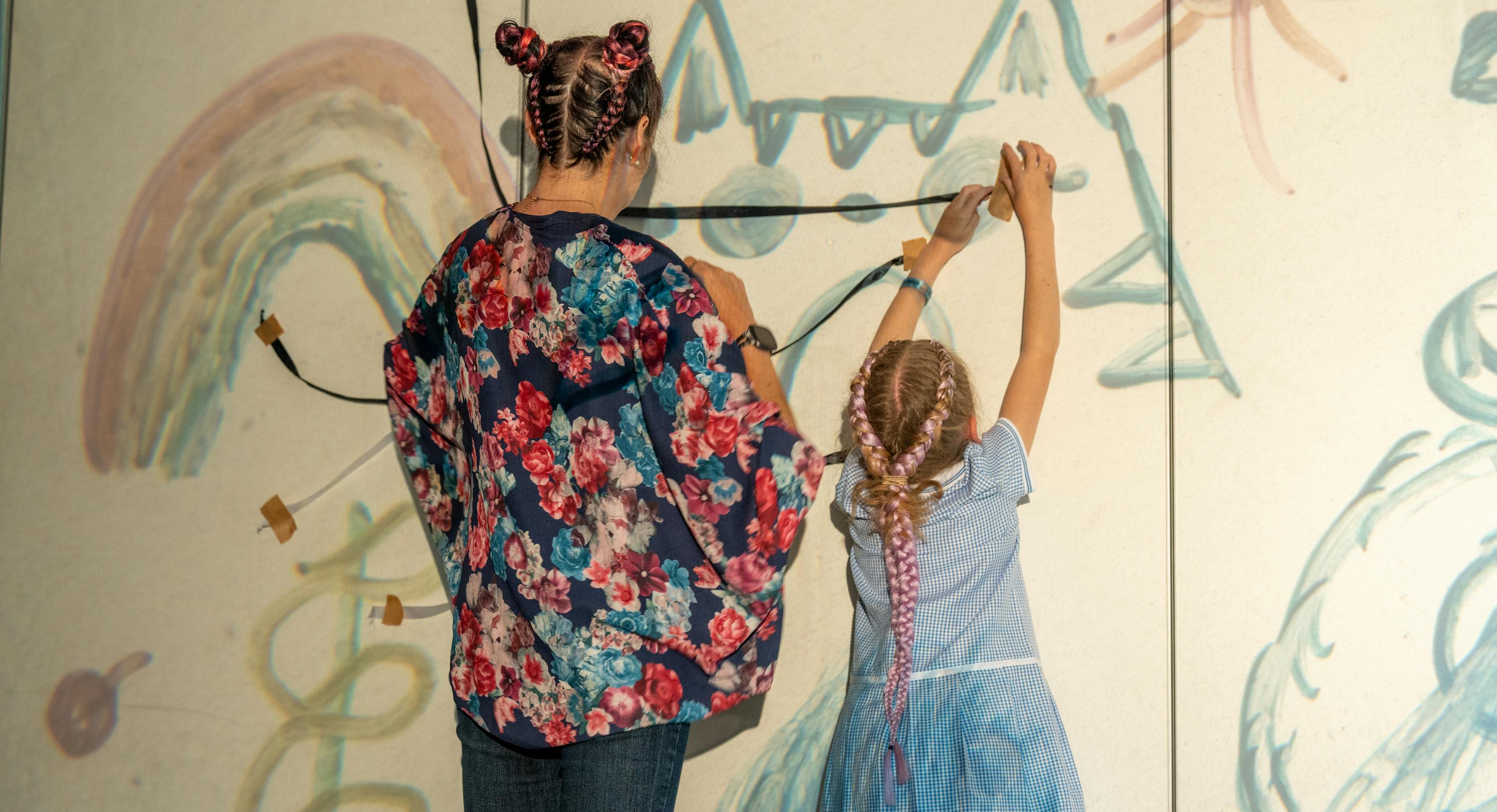 A young child and parent/carer are facing away from the camera. In front of them are illustrated drawings projected onto the wall and they making their own art piece within it with ribbon and tape