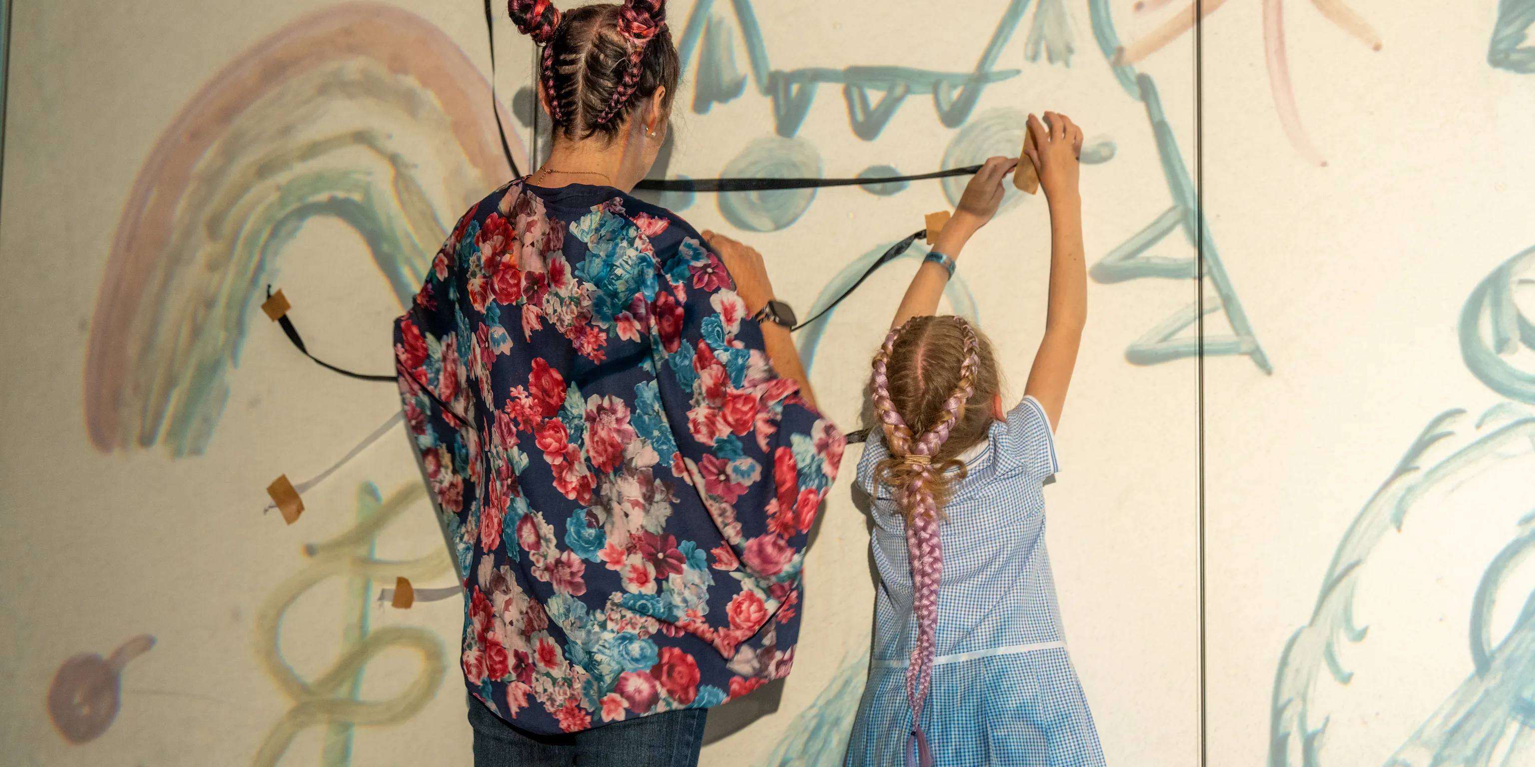 A young child and parent/carer are facing away from the camera. In front of them are illustrated drawings projected onto the wall and they making their own art piece within it with ribbon and tape