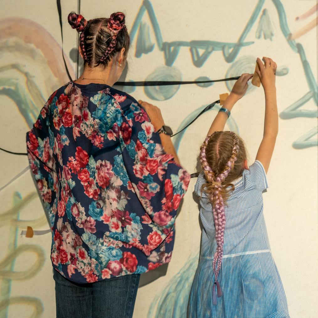 A young child and parent/carer are facing away from the camera. In front of them are illustrated drawings projected onto the wall and they making their own art piece within it with ribbon and tape
