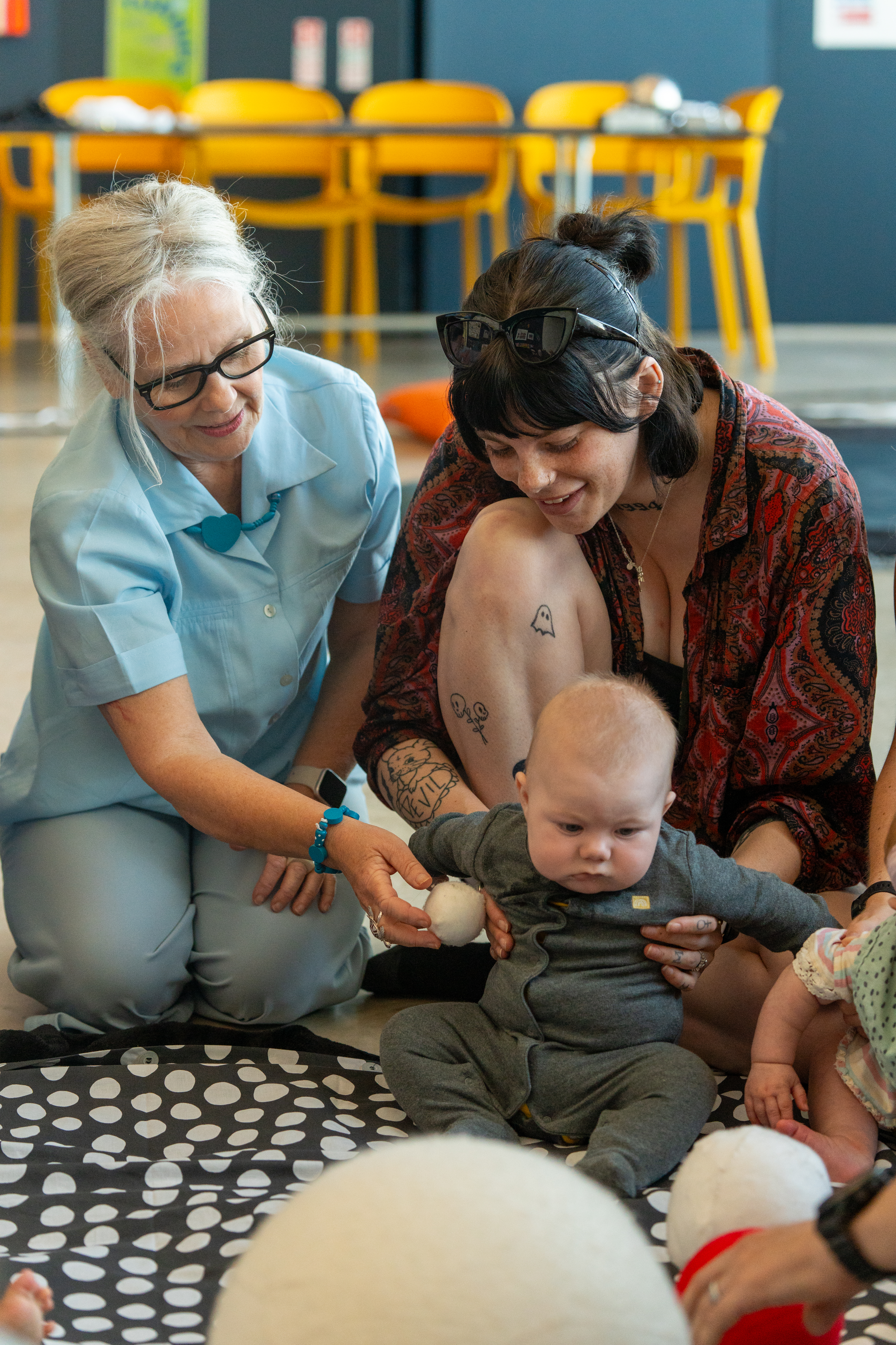 A baby, their parent/carer and Playground artists are sat on a black and white mat  during a Baby Playground session