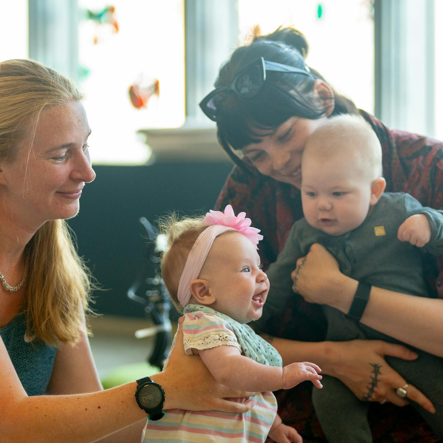 Two mums are holding their children during a Playground session. One of the babies is looking ahead and laughing, whilst the other is looking at them. Both parents are smiling.