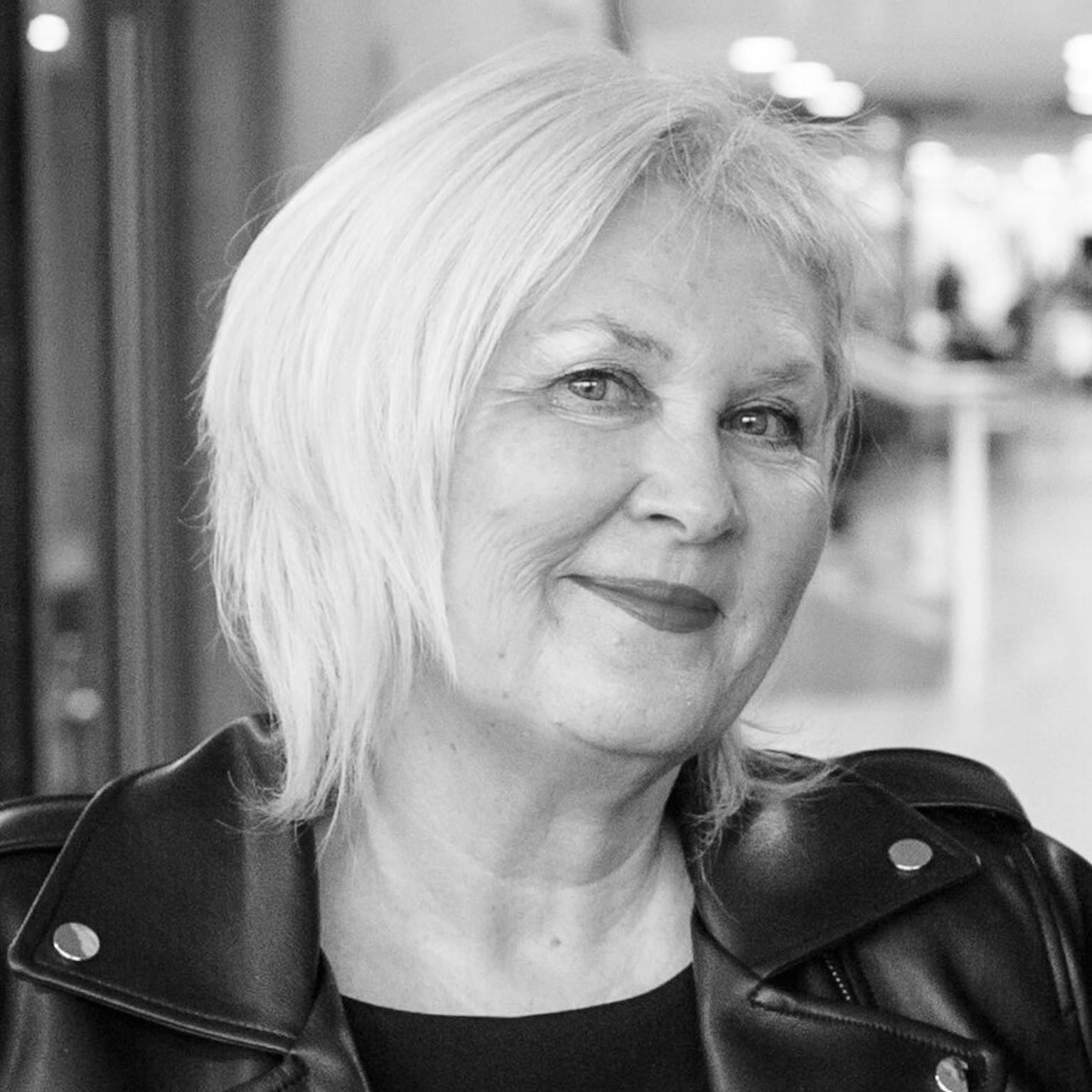 A black and white image of Liz. She has short blonde hair and is wearing a black leather jacket. She is looking to the camera and smiling.