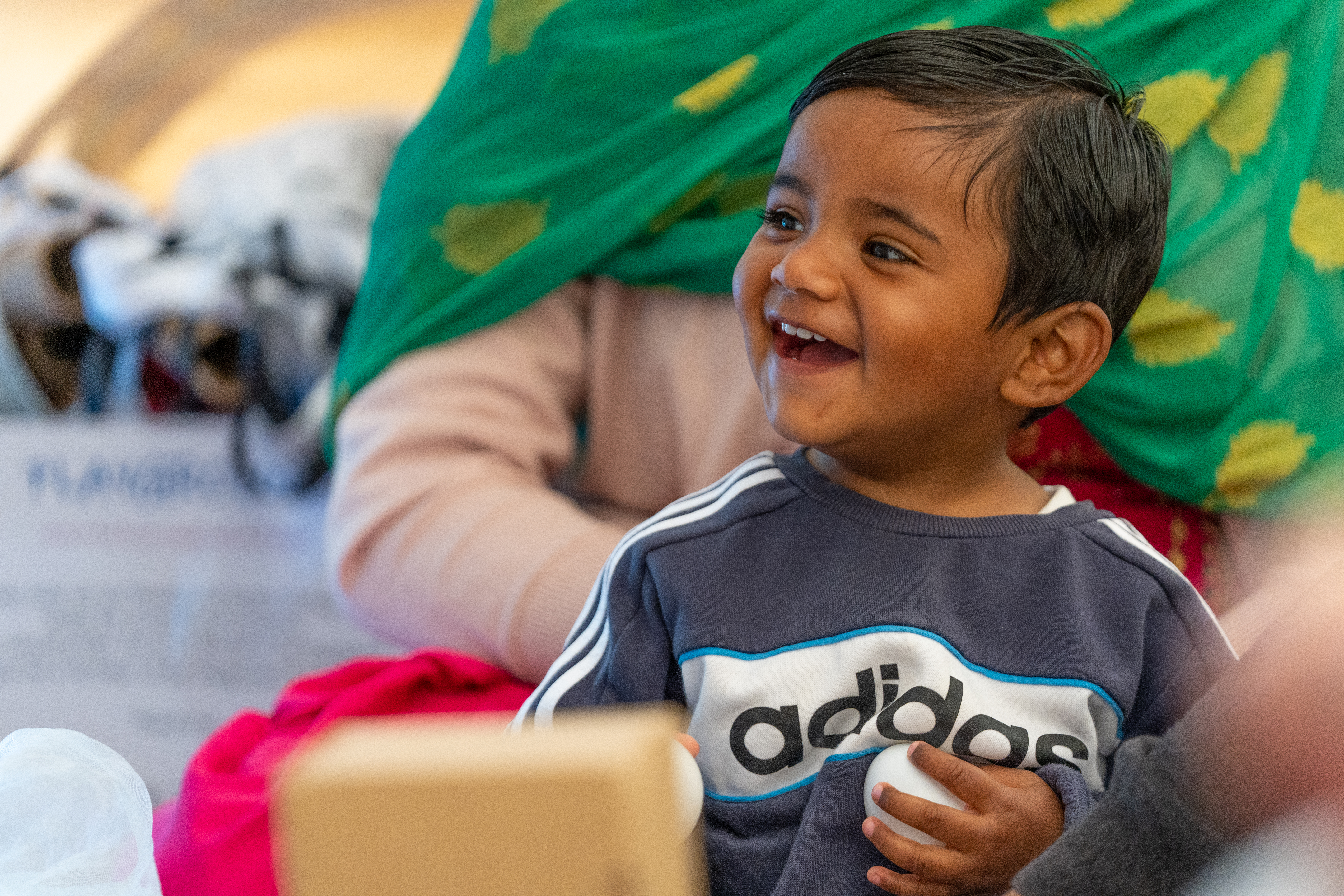 A young child is sat in front of their parent/carer and laughing. They are holding a small white ball in their hand.