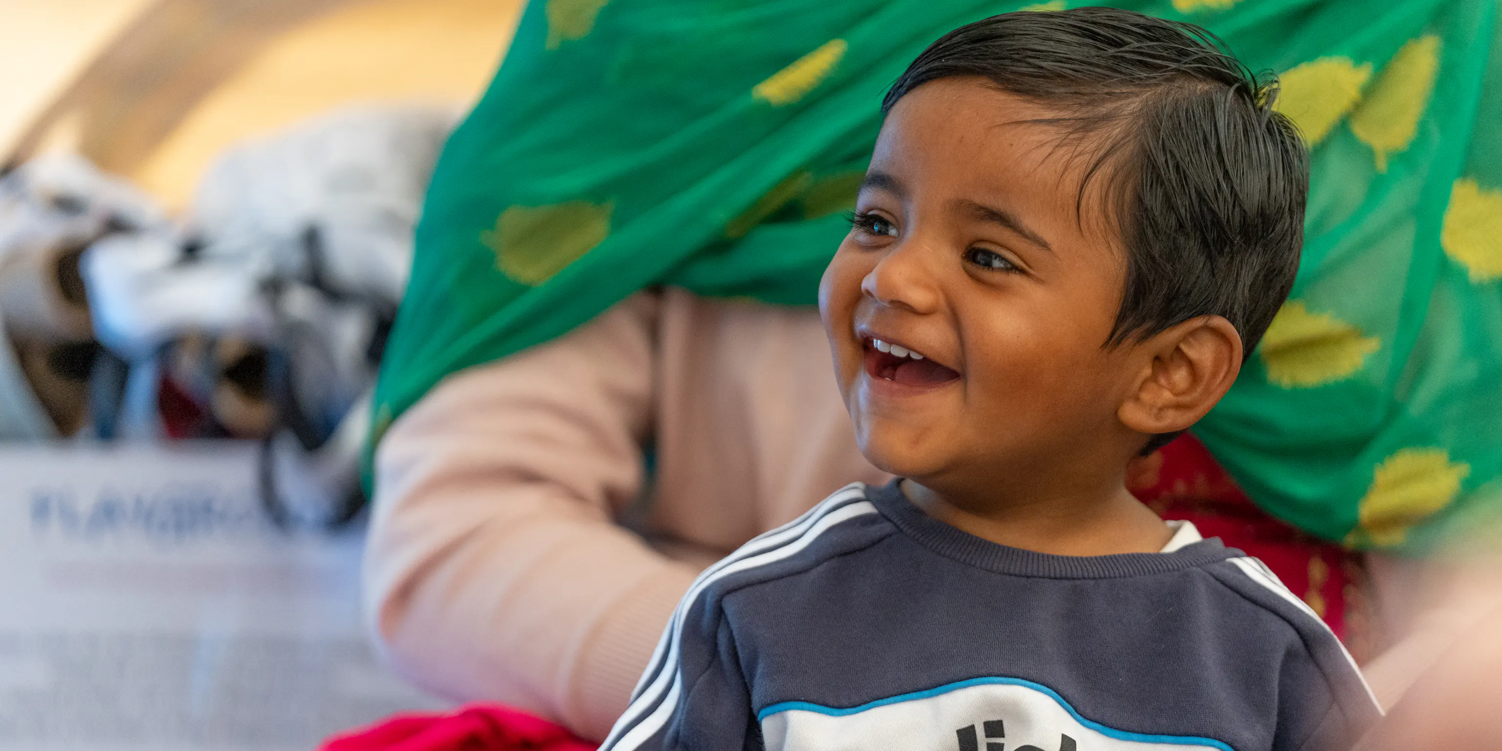 A young child is sat in front of their parent/carer and laughing. They are holding a small white ball in their hand.