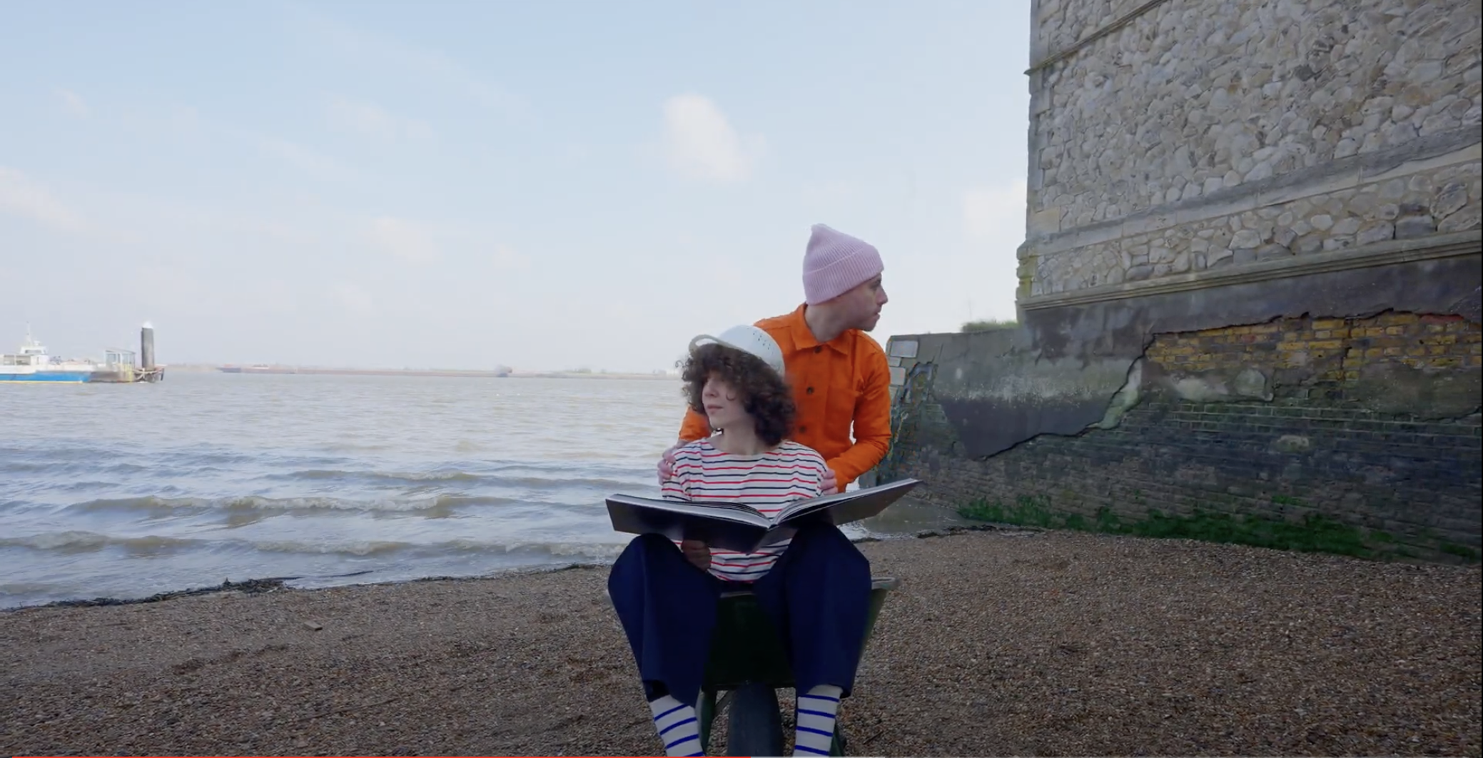 A person sat in a wheelbarrow with a book. Someone is pushing them and looking away.