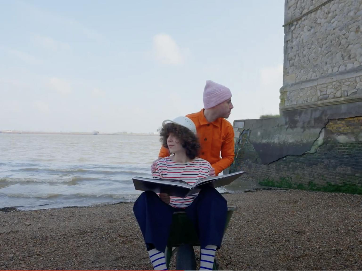A person sat in a wheelbarrow with a book. Someone is pushing them and looking away.