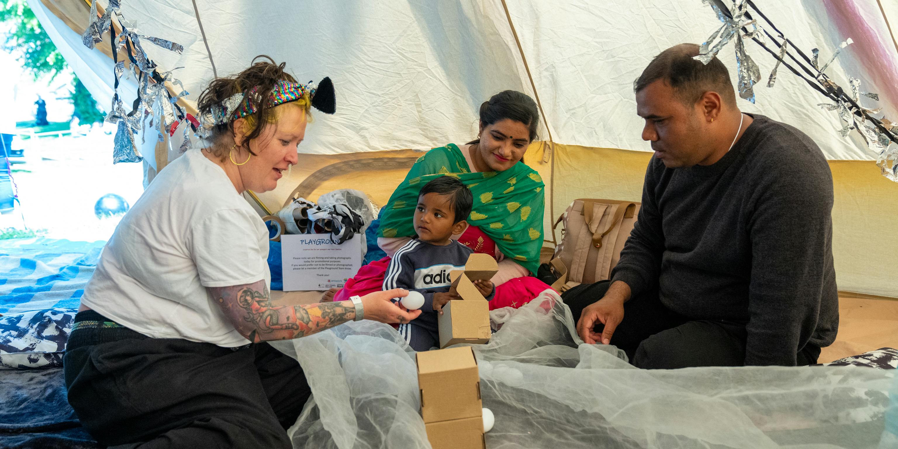A Playground artist is engaging with a young child and their parents/carers in a tipi tent at a festival. They are surrounded by boxes, white balls and netted materials