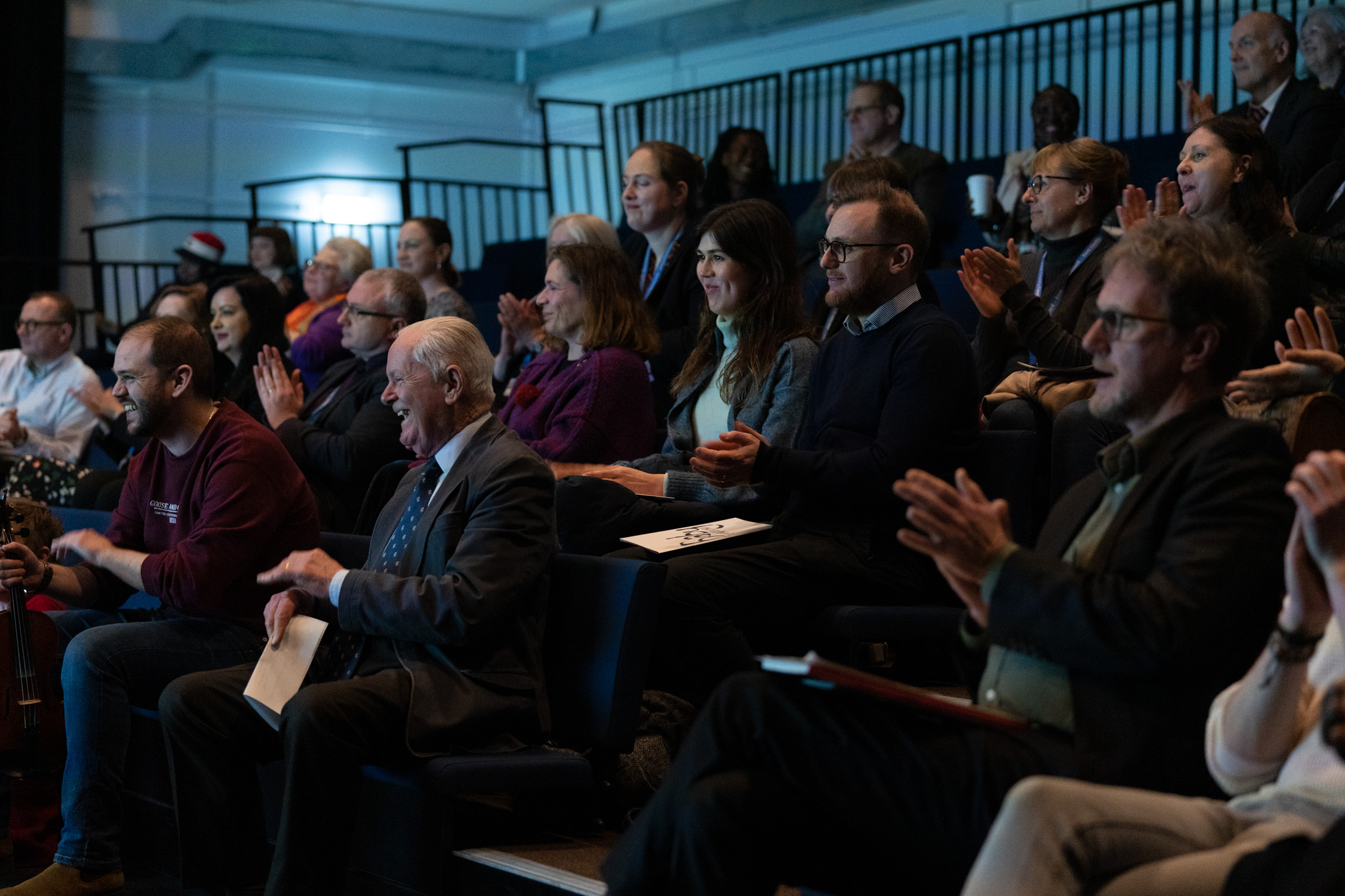 A group of people sat in a dark room together. Some people are clapping and others are smiling or laughing