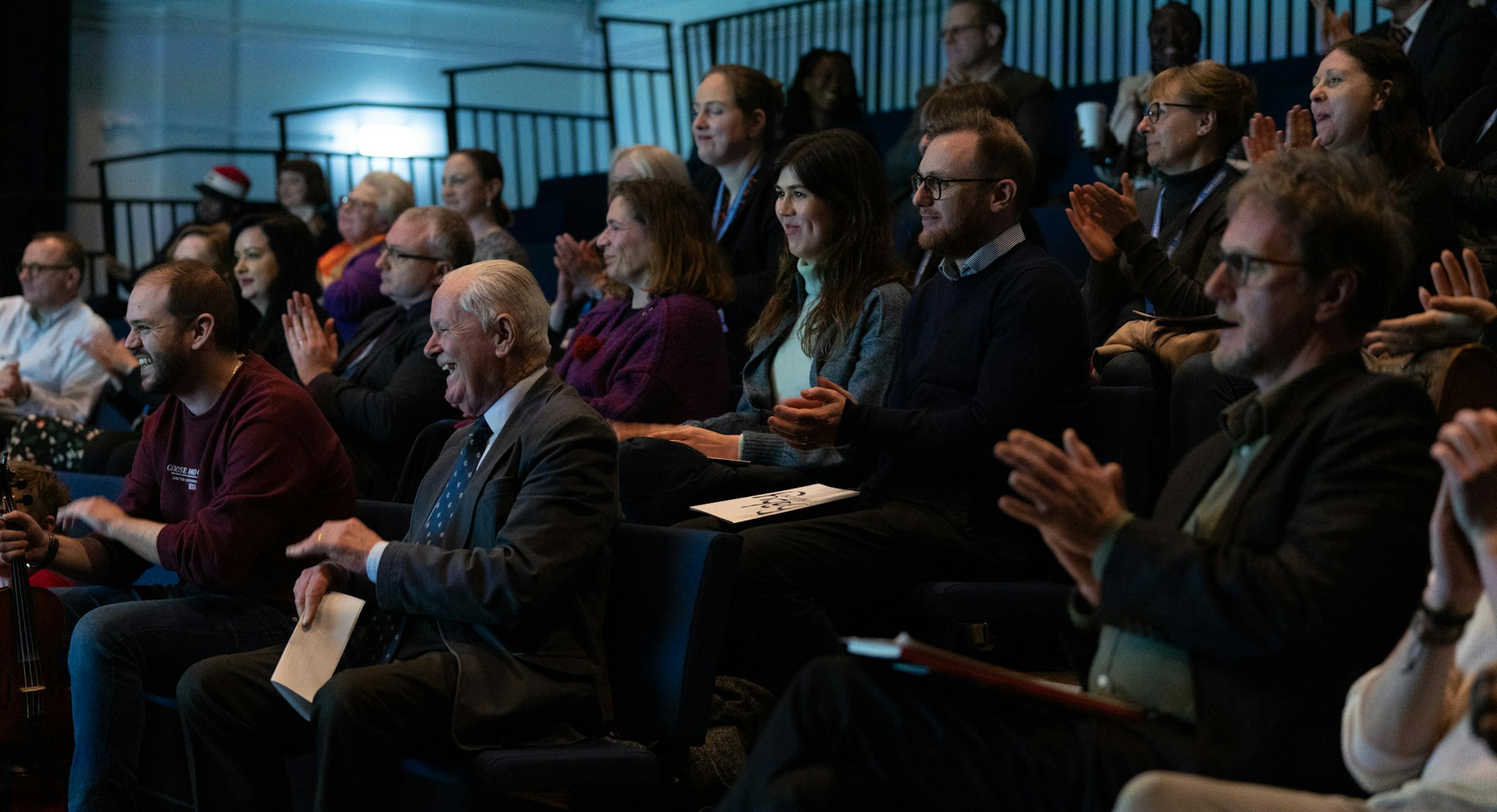 A group of people sat in a dark room together. Some people are clapping and others are smiling or laughing