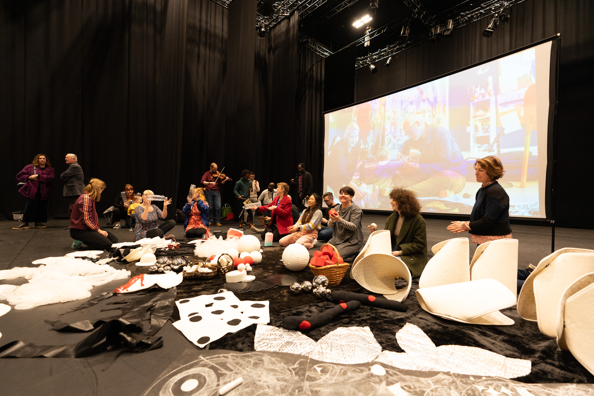 A group of artists are sat in a dark room and on the floor with a big screen behind them. They are all playing instruments or playing with materials. On the floor are a range of creative materials. 