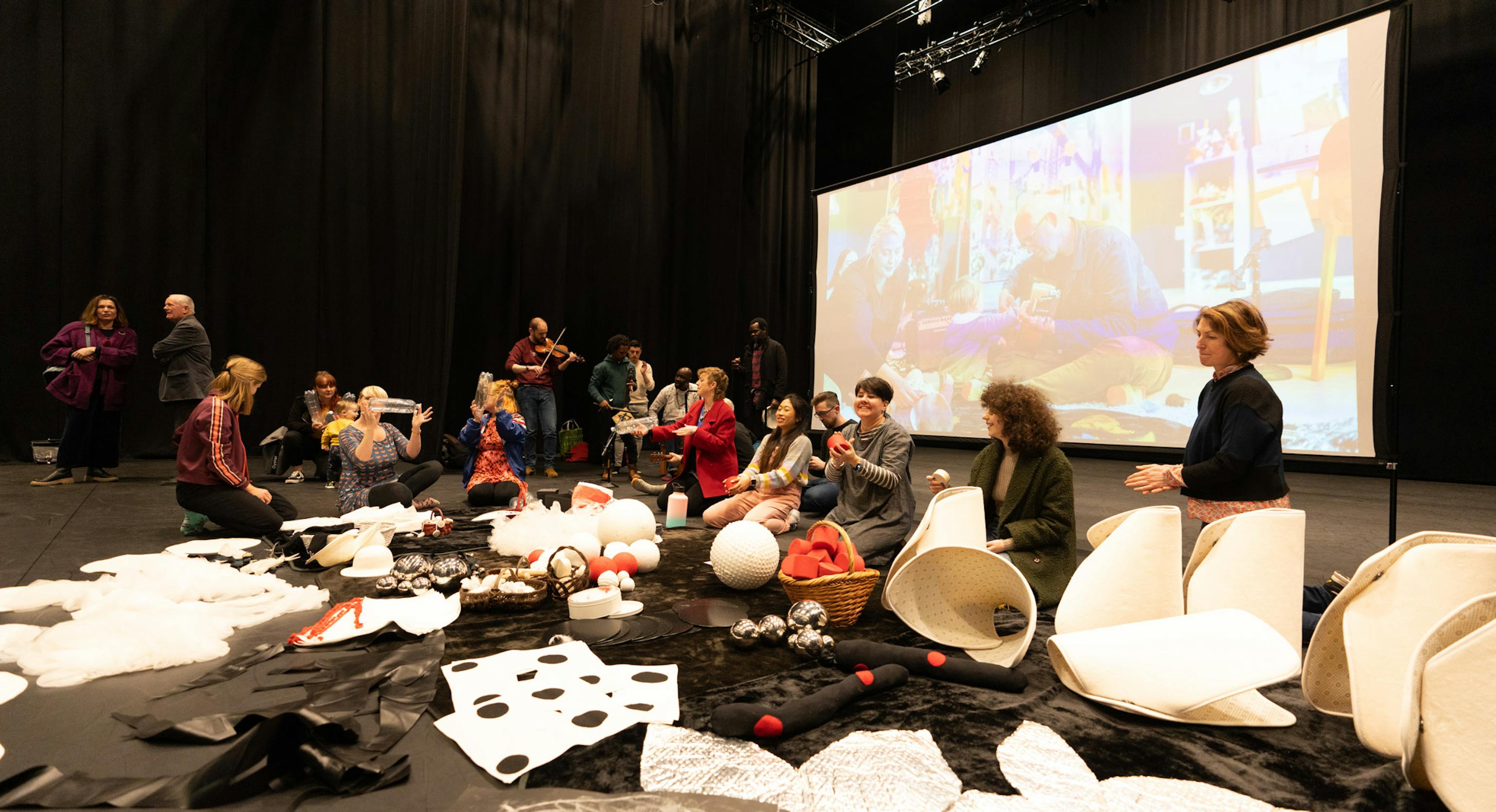 A group of artists are sat in a dark room and on the floor with a big screen behind them. They are all playing instruments or playing with materials. On the floor are a range of creative materials.