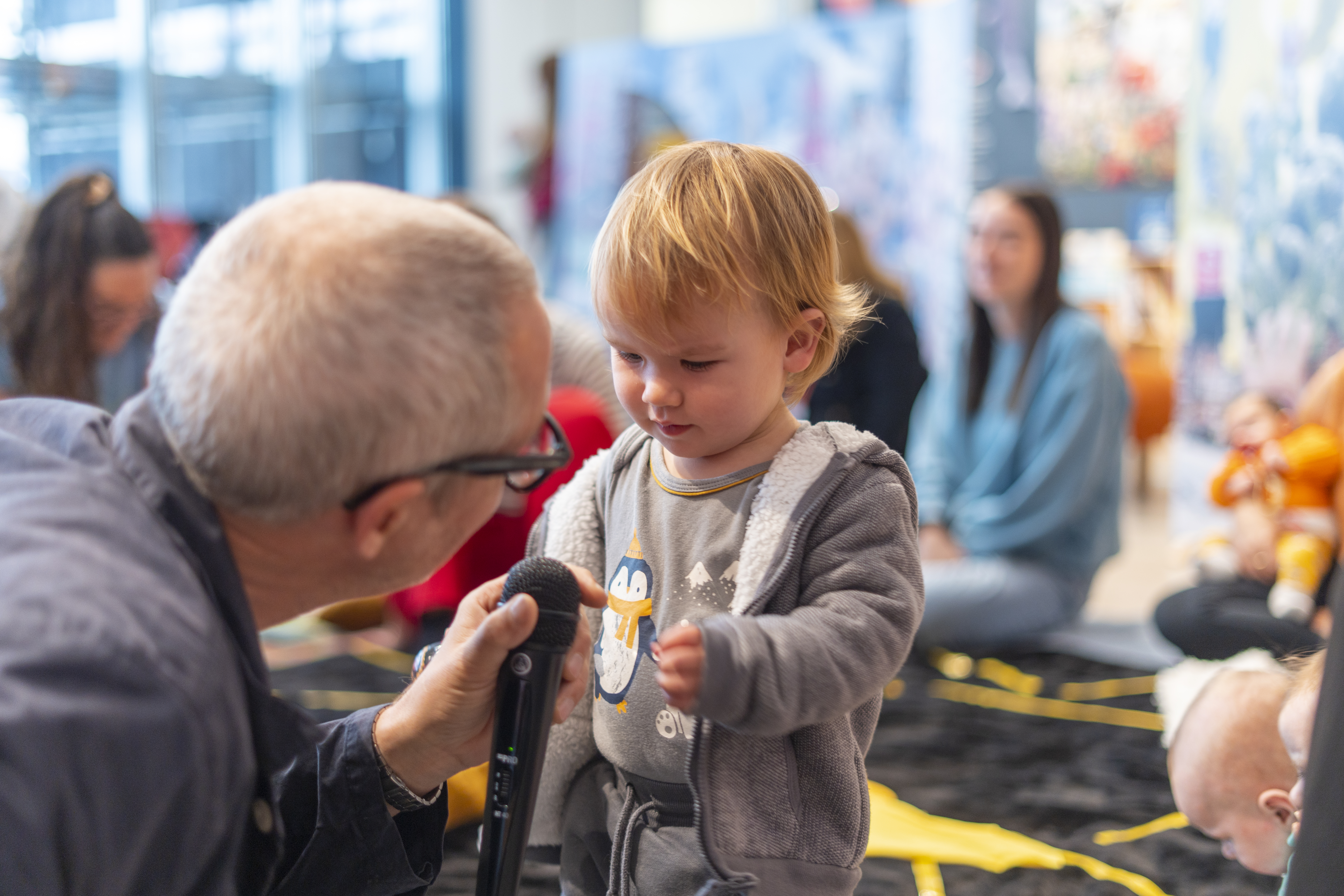 An artist is holding up a microphone to a baby who is reaching over for it 