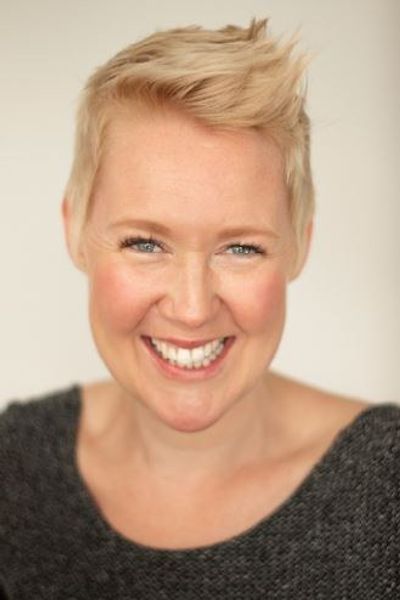 A woman with short blonde hair smiling at the camera. She is wearing a grey jumper. 