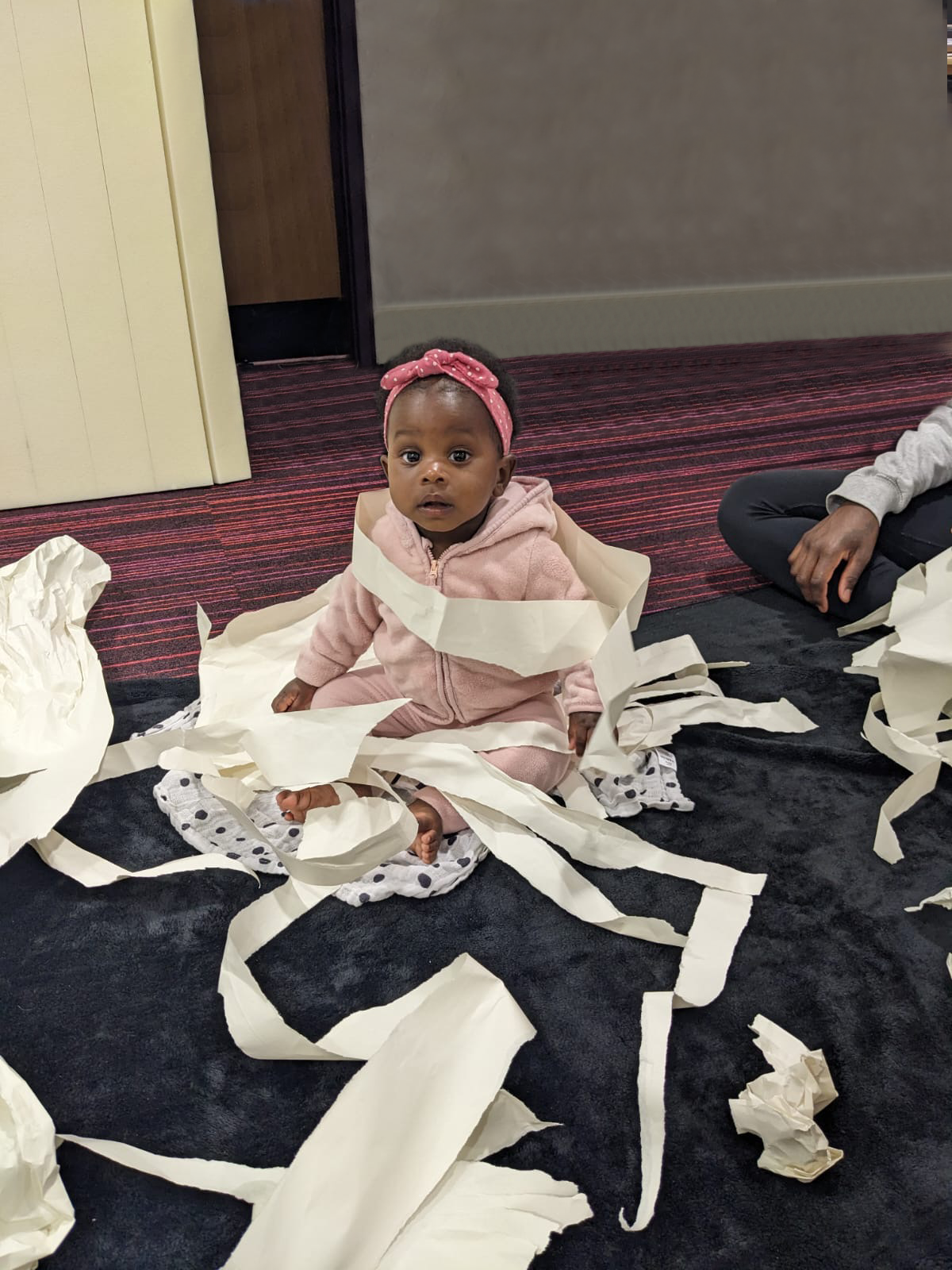 A baby is sat on a textured may and looking at the camera. They are surrounded by shreds of white paper on and around them.