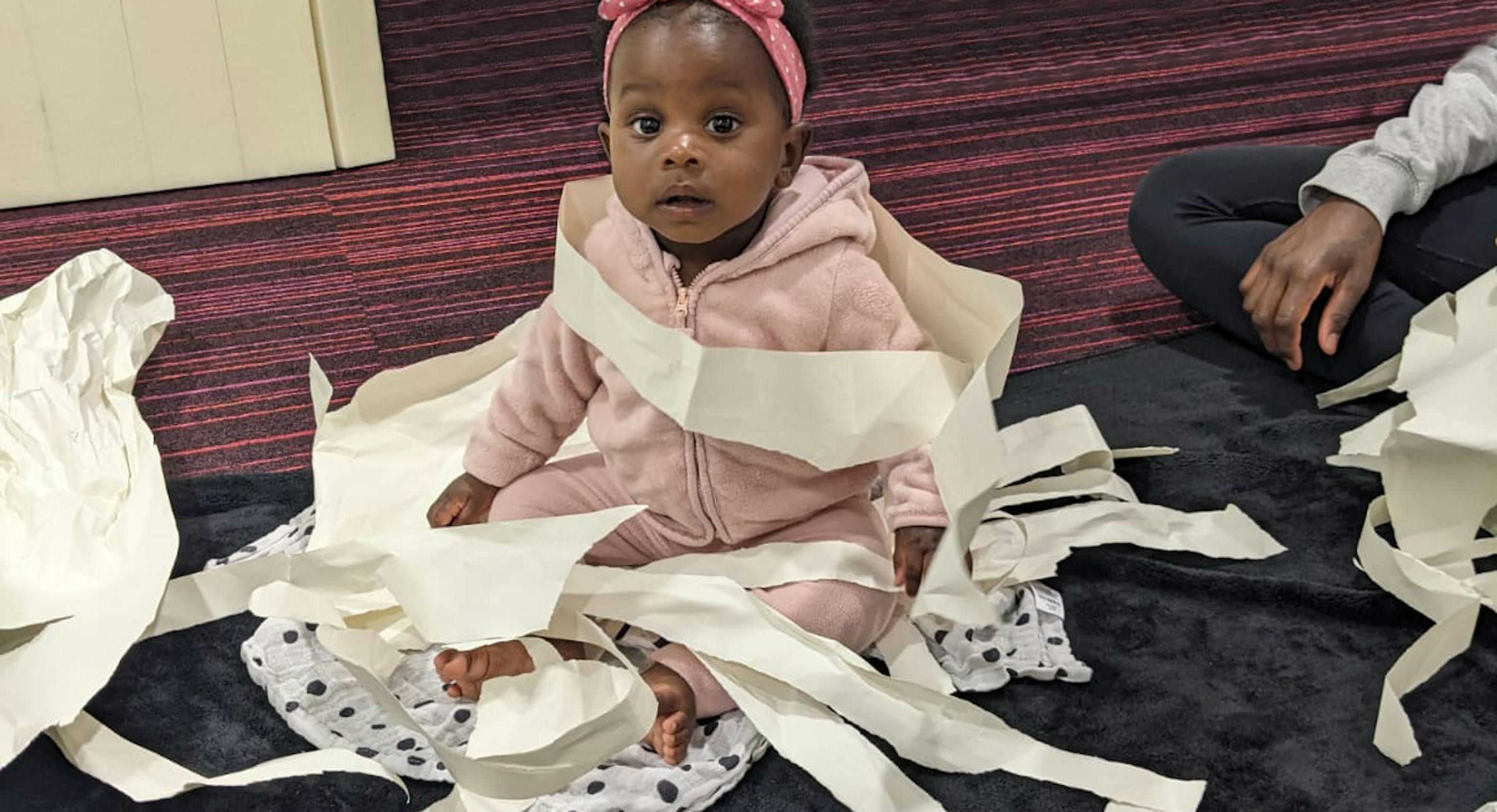 A baby is sat on a textured may and looking at the camera. They are surrounded by shreds of white paper on and around them.