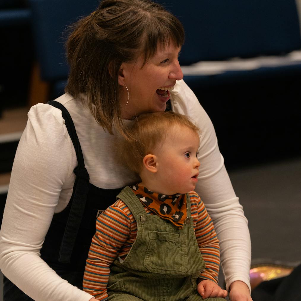 A young child is sitting on their parent/carer's lap and looking at something around them. Their parent/carer is laughing and looking in the same direction as the young child.