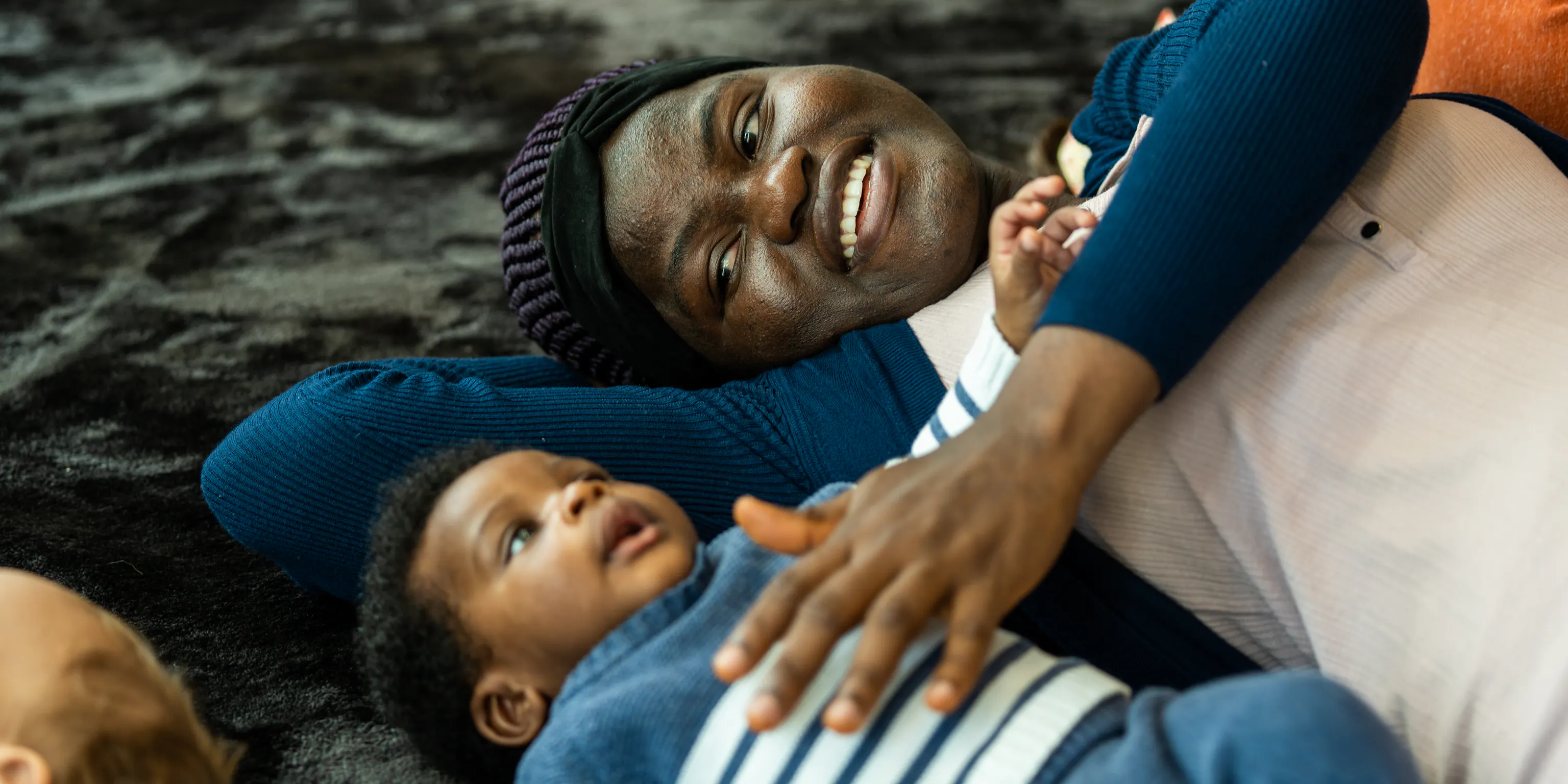 A baby is lying on a textured mat and looking up at the ceiling. Their parent/carer is lying next to them and touching their chest.