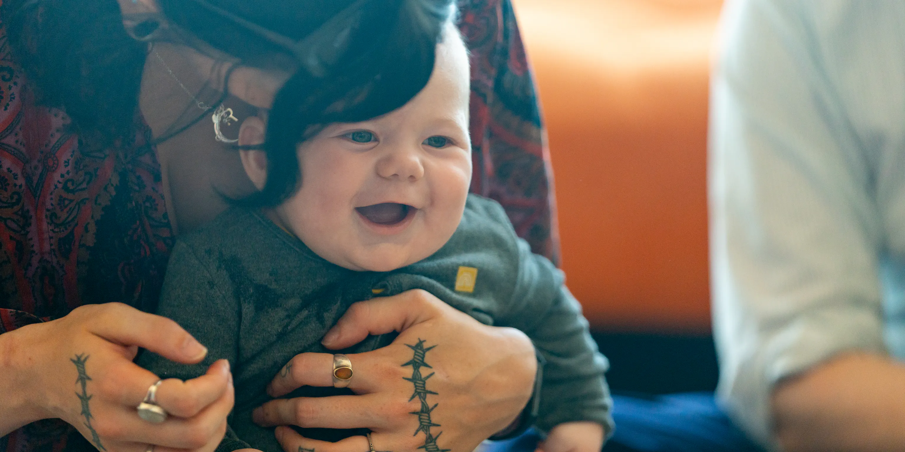 A parent/carer is sat with a baby and hugging them. The baby is laughing and looking at something in front of them