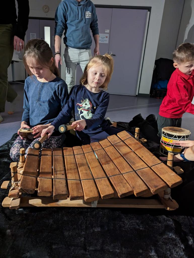 Two young children playing with musical instruments. The one on the left is playing with a mbira/kalimba instrument and the other a xylophone/marimba instrument.