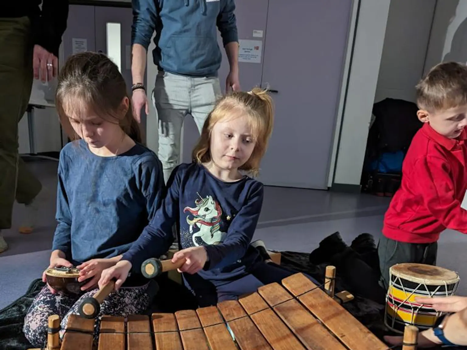 Two young children playing with musical instruments. The one on the left is playing with a mbira/kalimba instrument and the other a xylophone/marimba instrument.