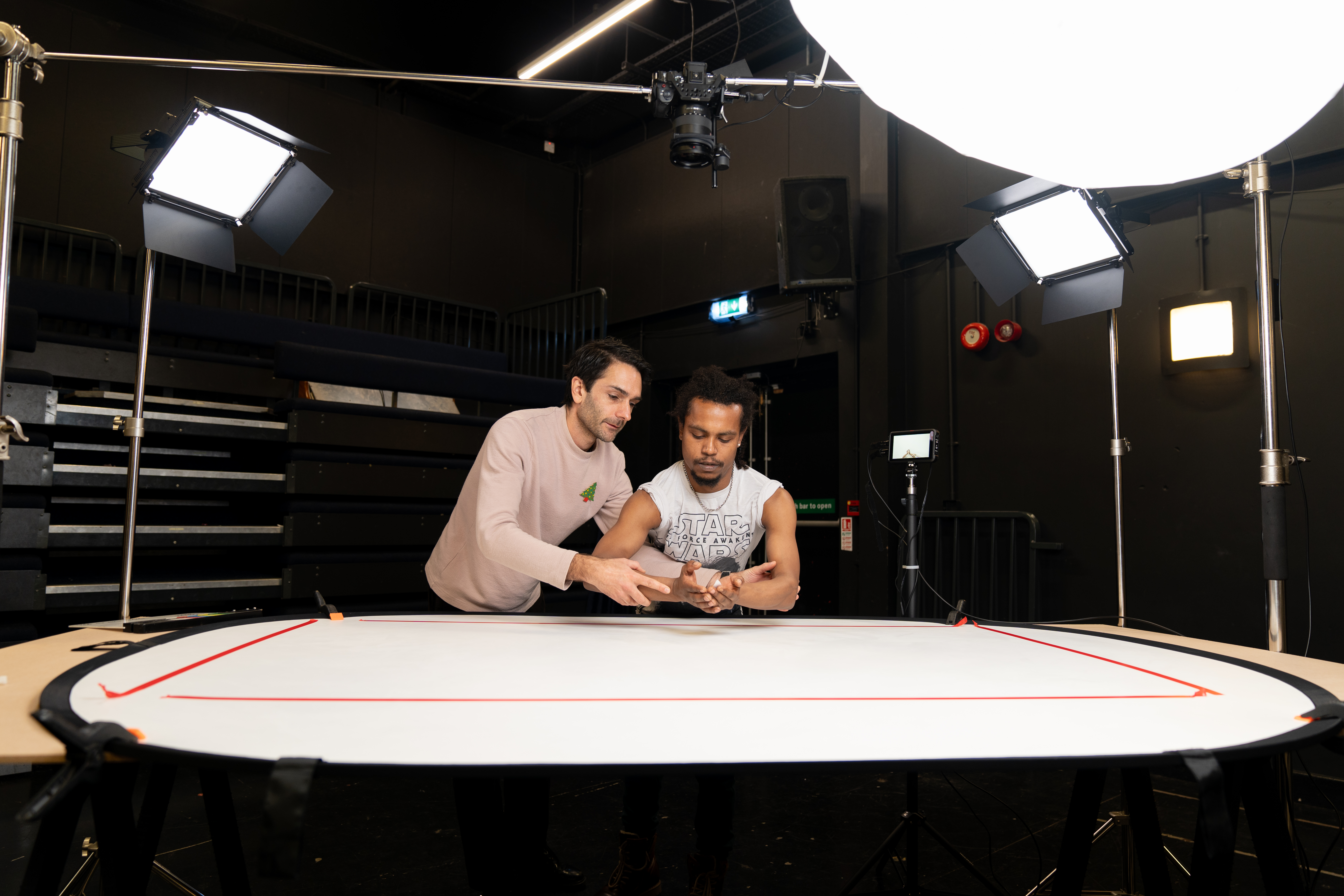 Two artists are stood next to each other, holding and crossing their arms in preparation of shooting a video. Around them are bright lights used for filming