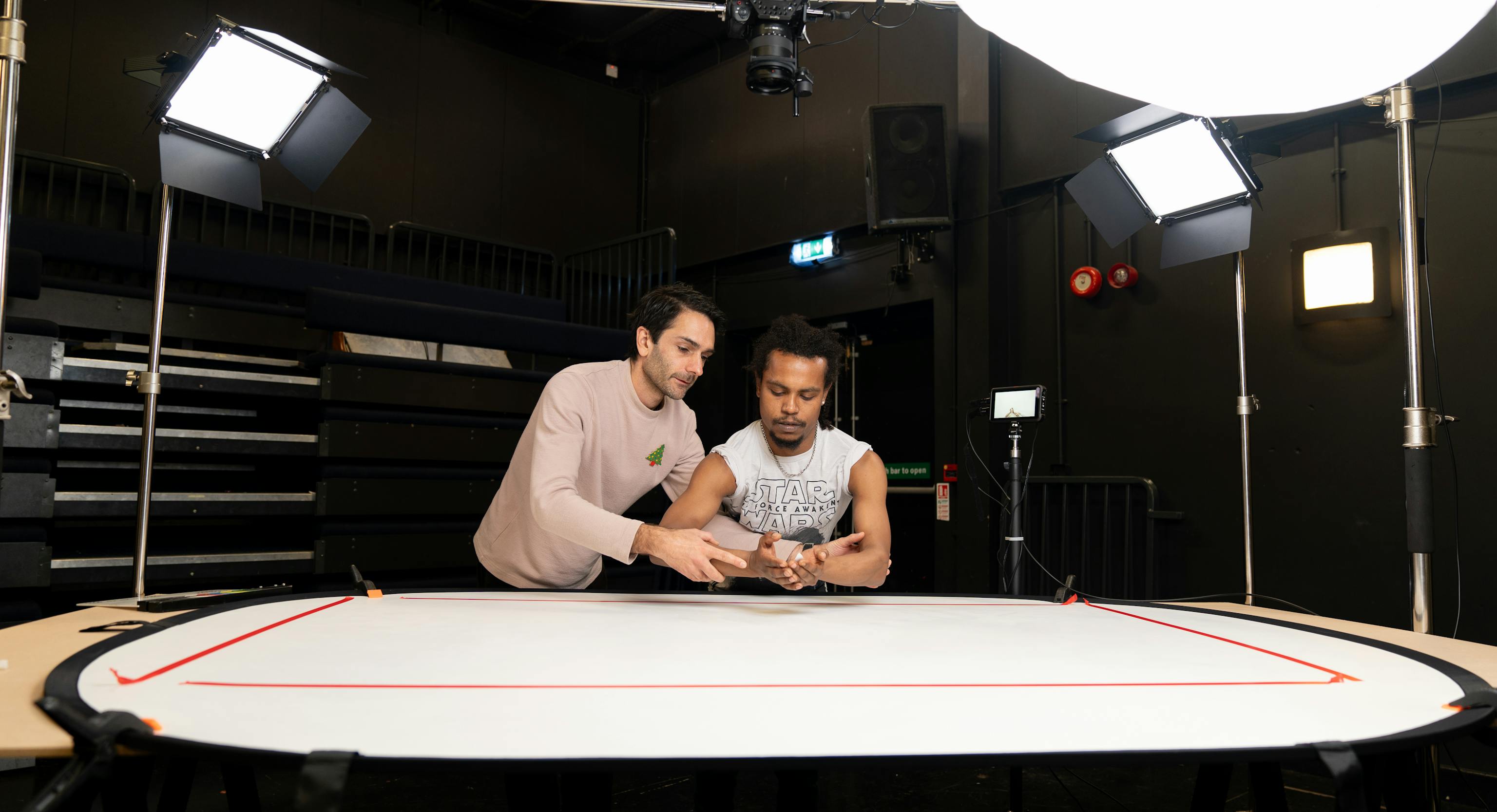 Two artists are stood next to each other, holding and crossing their arms in preparation of shooting a video. Around them are bright lights used for filming