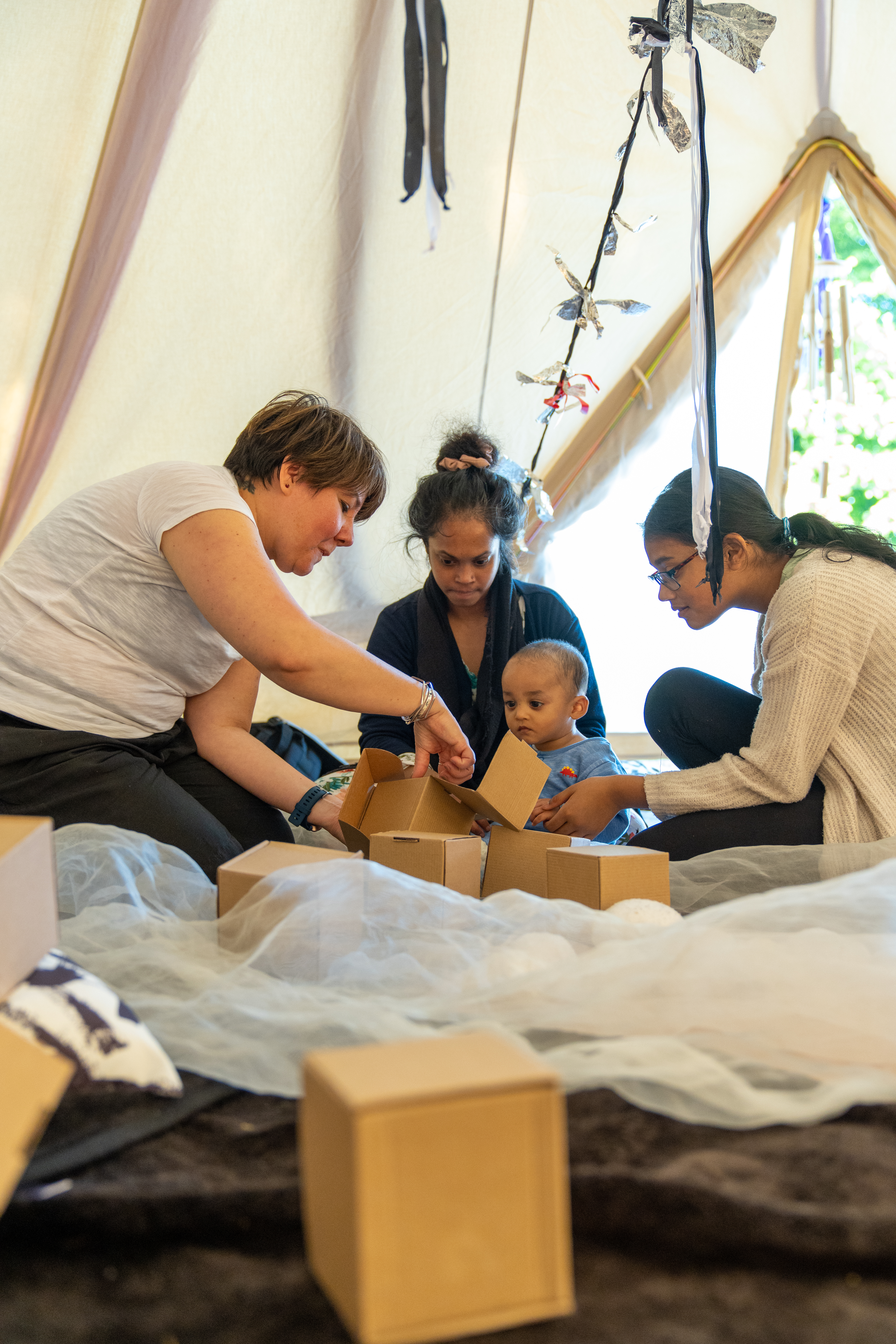 A small group with a baby, their parent/carers and Playground artist are sat in a tipi together with boxes and white netting material surrounding them