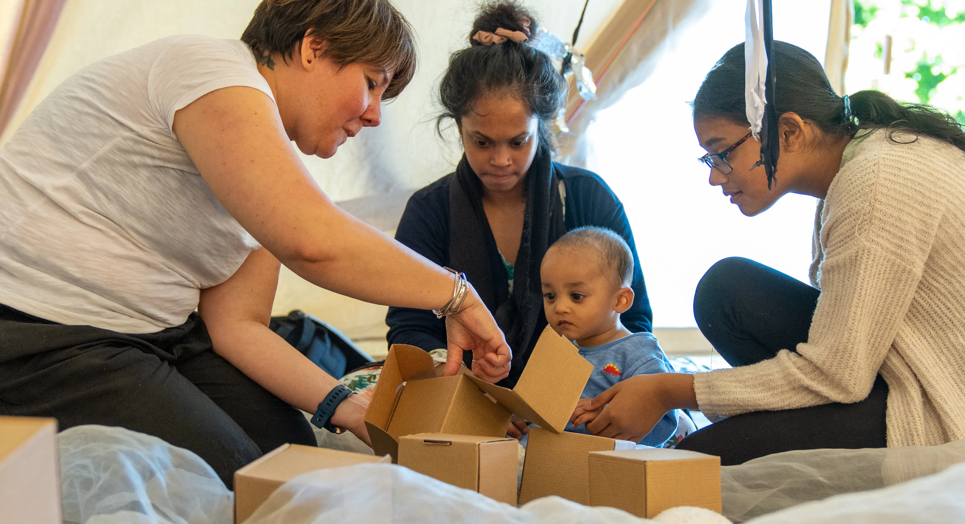 A small group with a baby, their parent/carers and Playground artist are sat in a tipi together with boxes and white netting material surrounding them