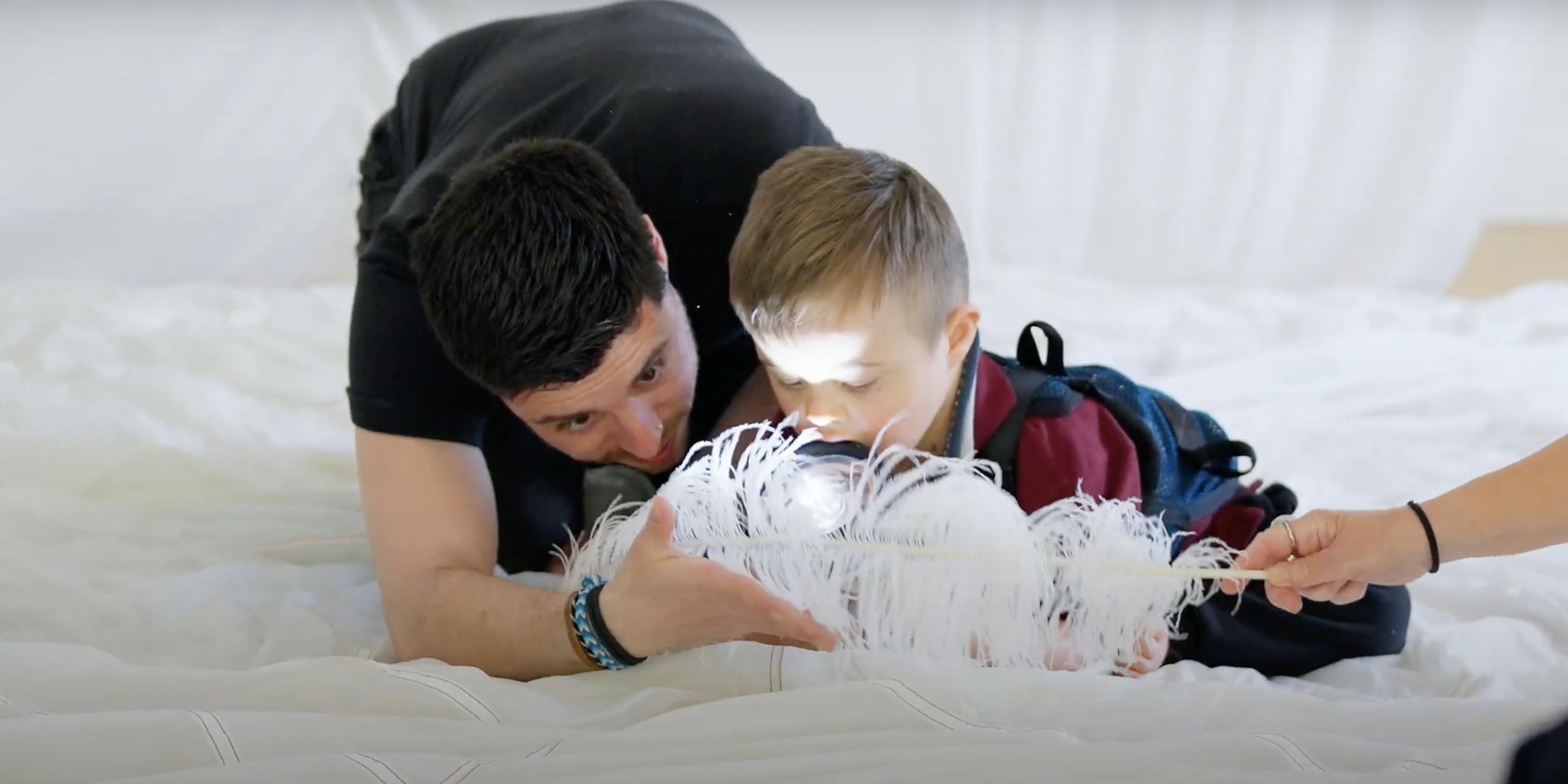 A young child and Playground artist are on a white piece of material together. They are playing with a feather and torch.
