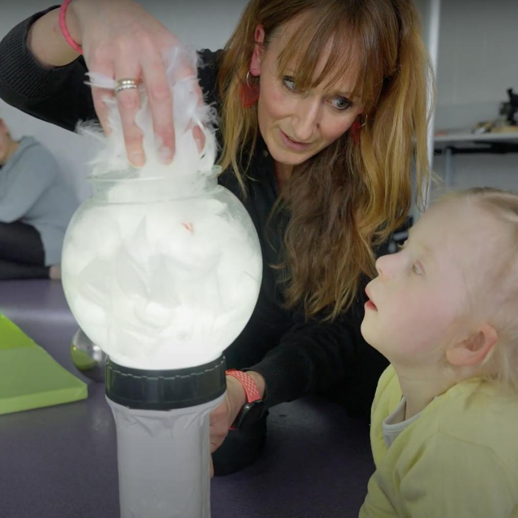 A Playground artist and young girl are engaging with a lit torch that has a bowl and white feathers in it. The young girl is looking at the artists hand that is holding the feathers. In the background is the child's mother
