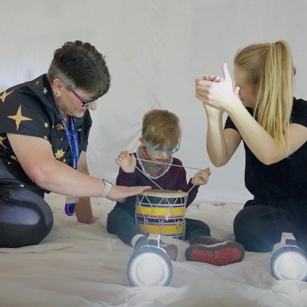 A support worker, young child and Playground artist are sat together playing with instruments and torch lights. The artist to the right is clapping their hands