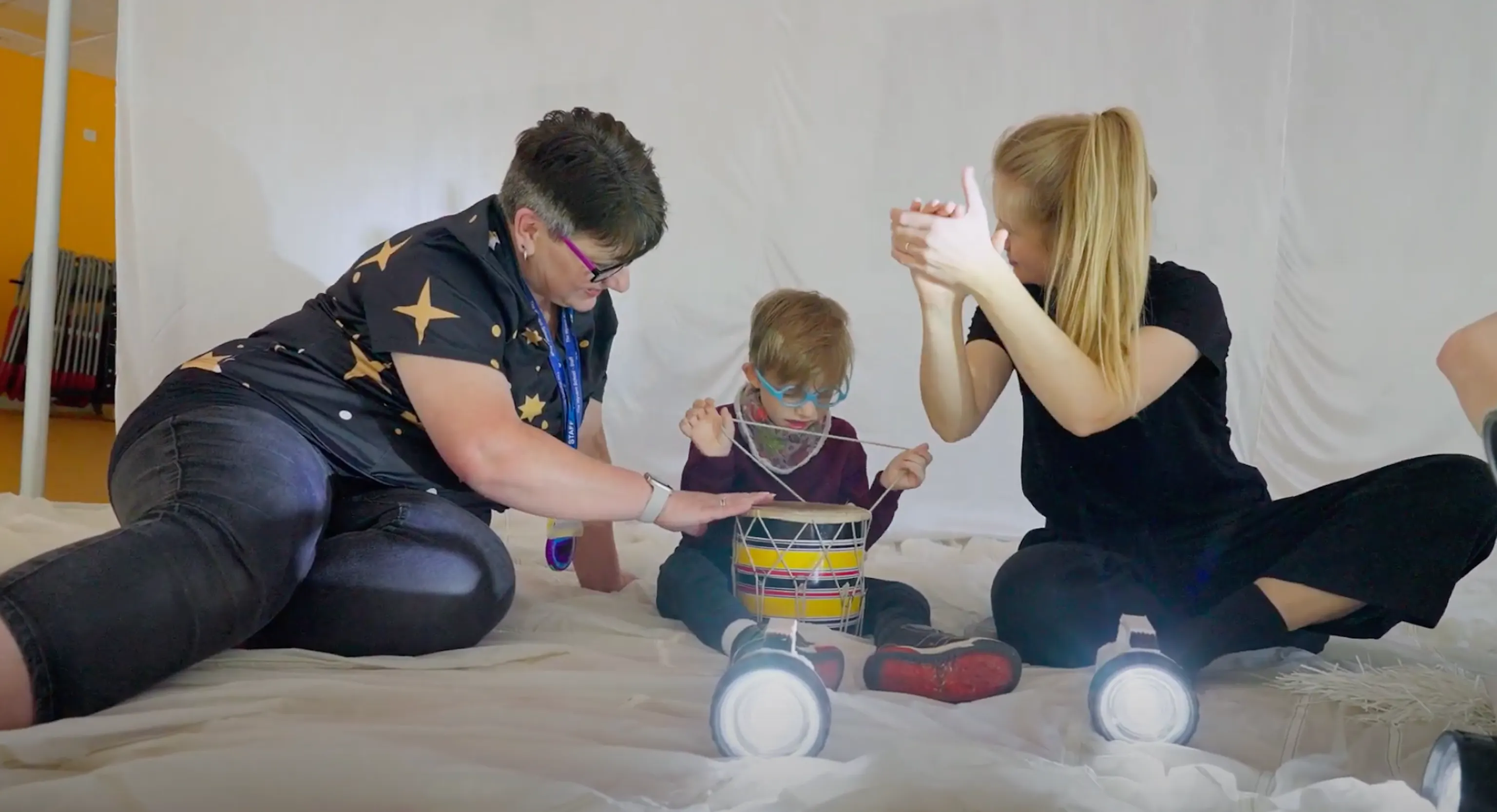 A support worker, young child and Playground artist are sat together playing with instruments and torch lights. The artist to the right is clapping their hands