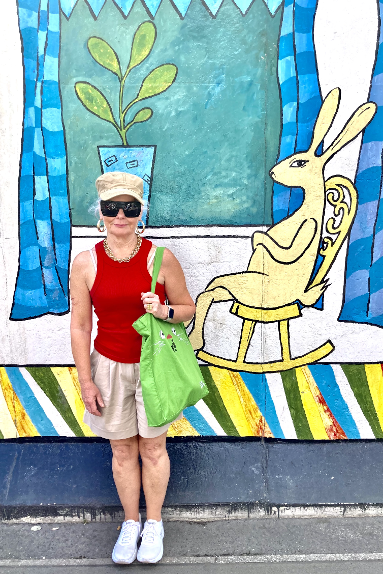 An artist wearing sunglasses and a hat stood in front of a colourful background. The background includes a plant pot and rabbit to the right of the screen.
