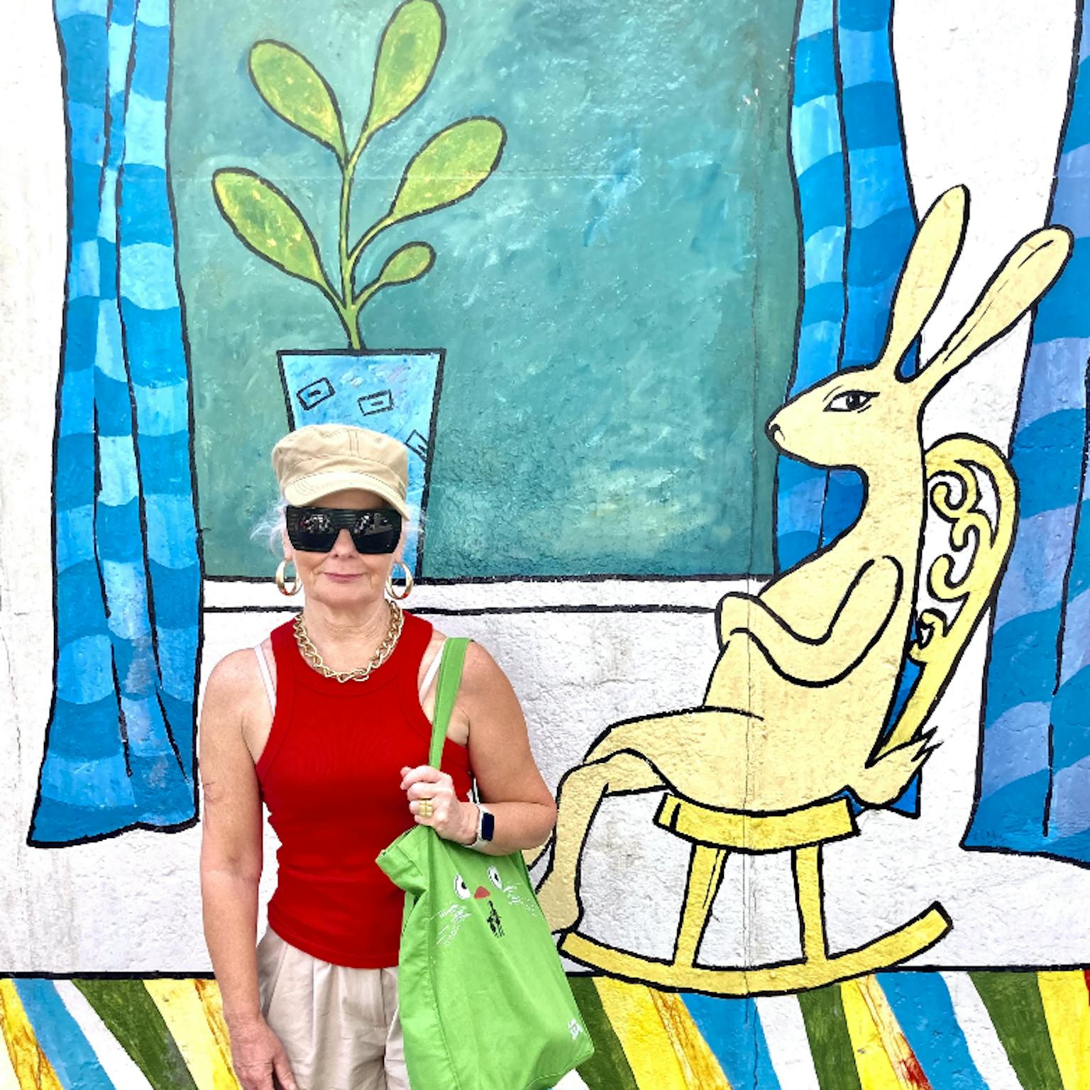 An artist wearing sunglasses and a hat stood in front of a colourful background. The background includes a plant pot and rabbit to the right of the screen.