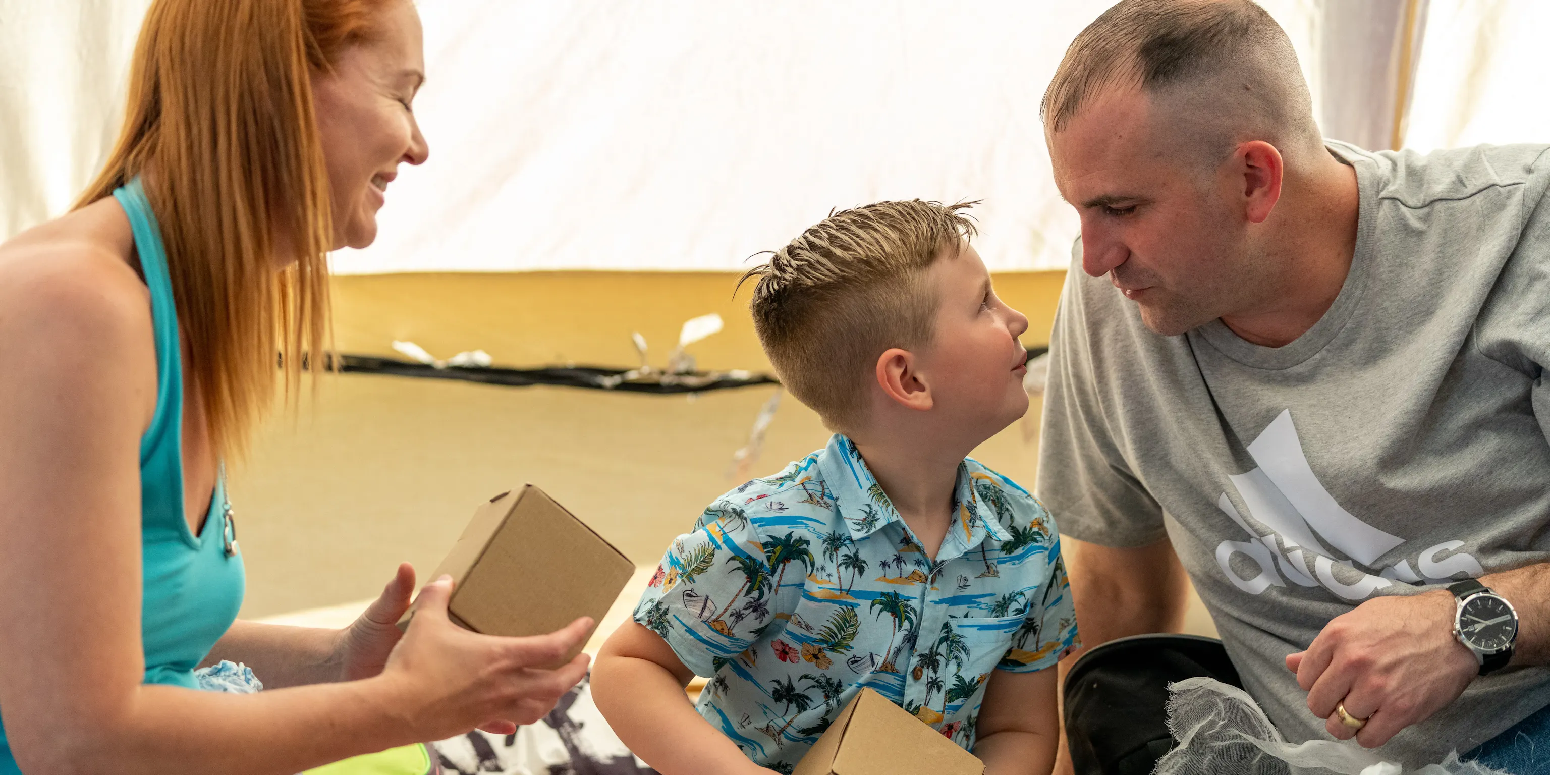 A young child sat in-between their parent/carers. They are holding boxes and looking at each other