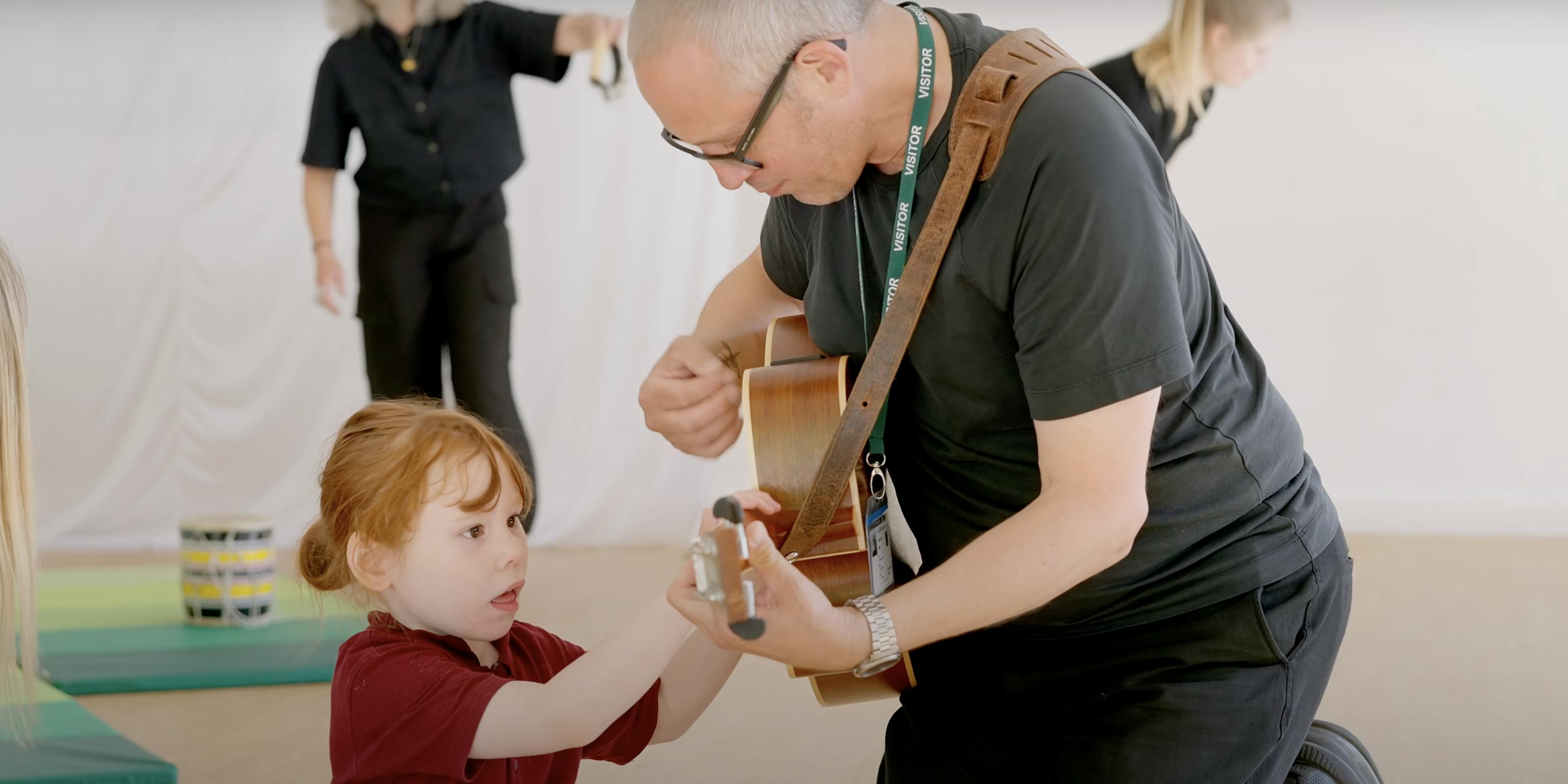 An artist playing a guitar. The child next to them is looking and touching the guitar