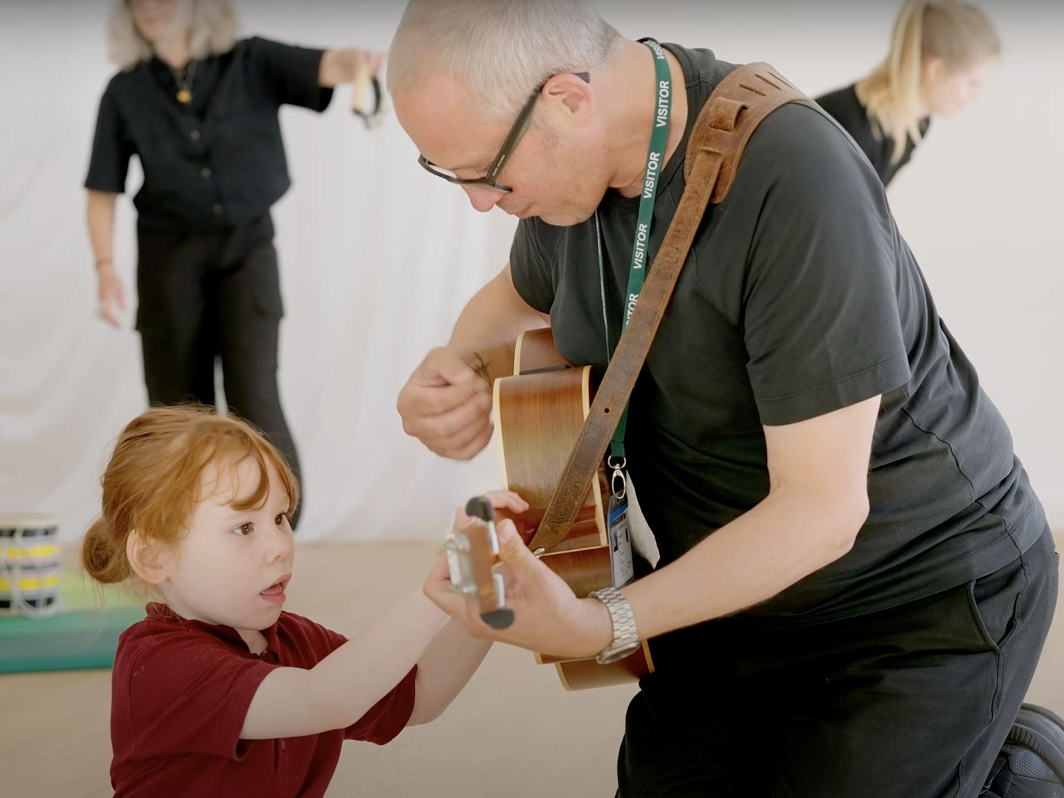 An artist playing a guitar. The child next to them is looking and touching the guitar