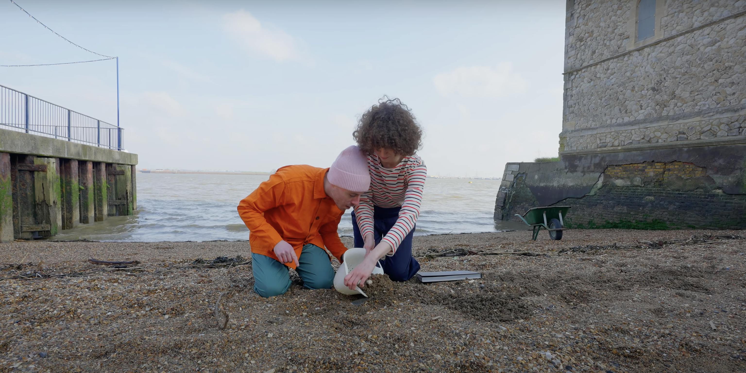 Two people on the beach. They are using a colander to dig up the sand to see what they will uncover