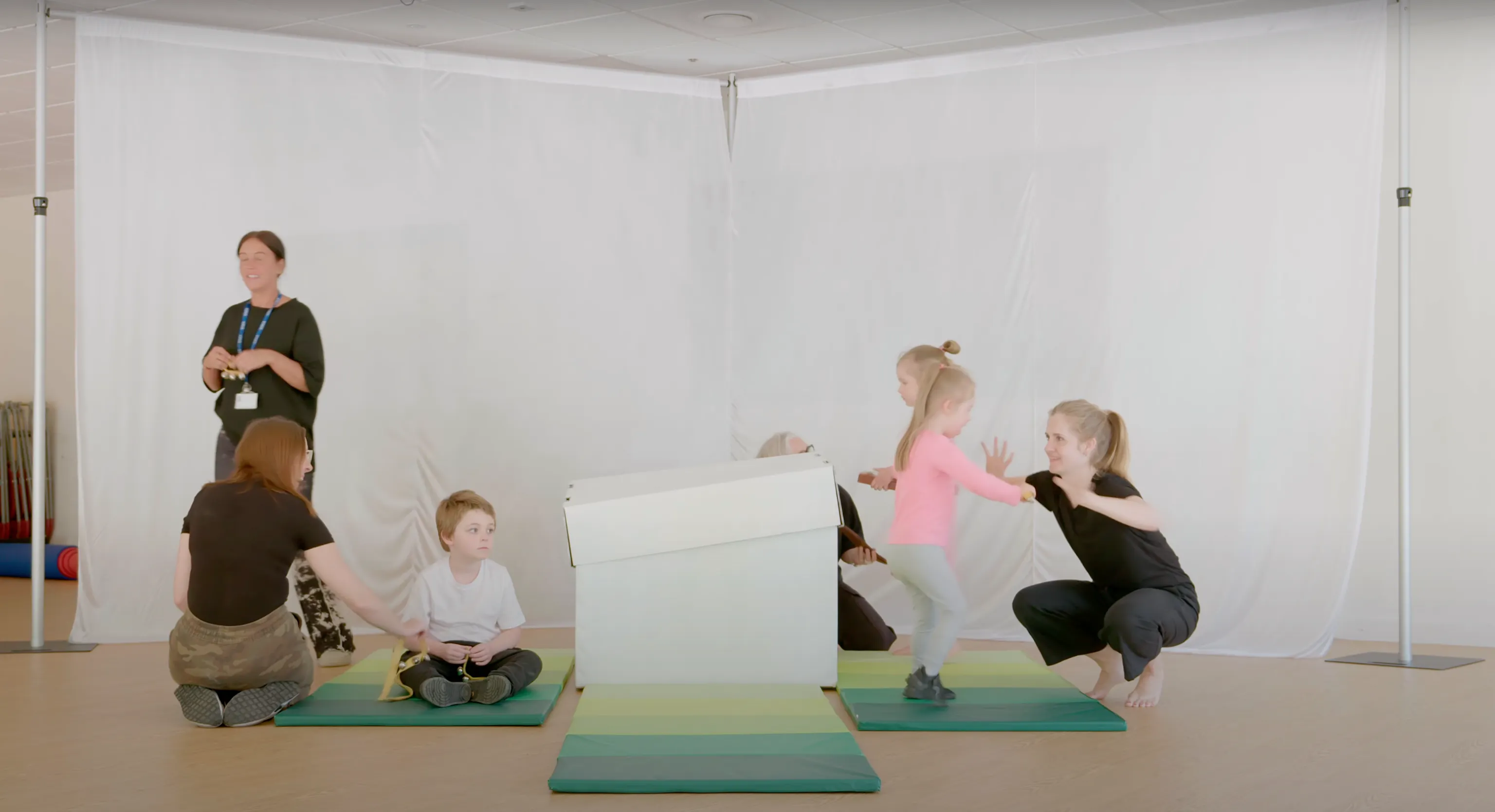 A Playground session with a group of young children, artists and their carers all engaging with each other on green mats. In the middle is a big white box