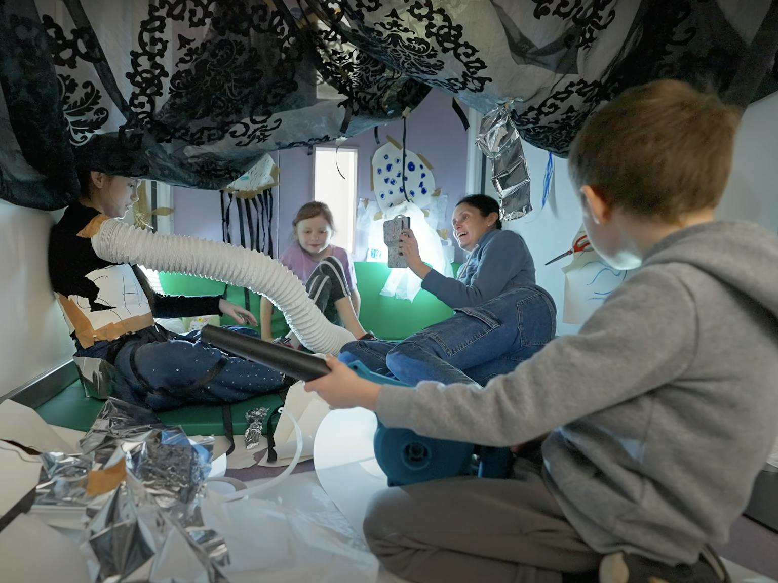 A family together during a Family Playground session. There is a black net above them and a lot of foil and paper material on the floor