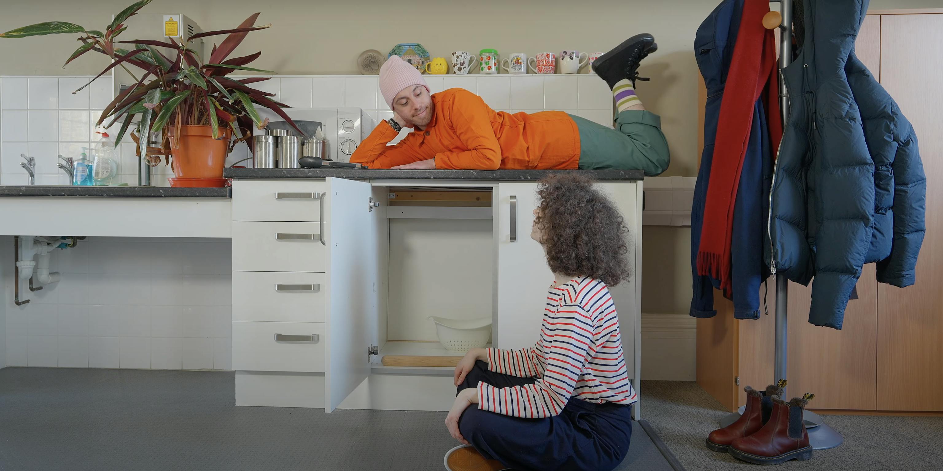 A person lying on a kitchen counter is smiling at the person sat on a floor with their legs crossed. They are surrounded by kitchen utensils and to the right is a coat rack