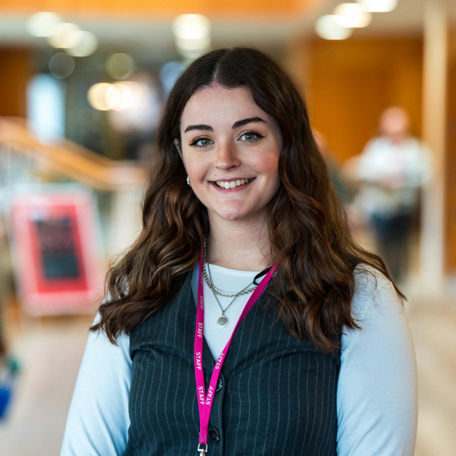 a young white woman with mid-length brown curly hair and green eyes, wearing a white long sleeve t-shirt and a black waistcoast smiling at the camera