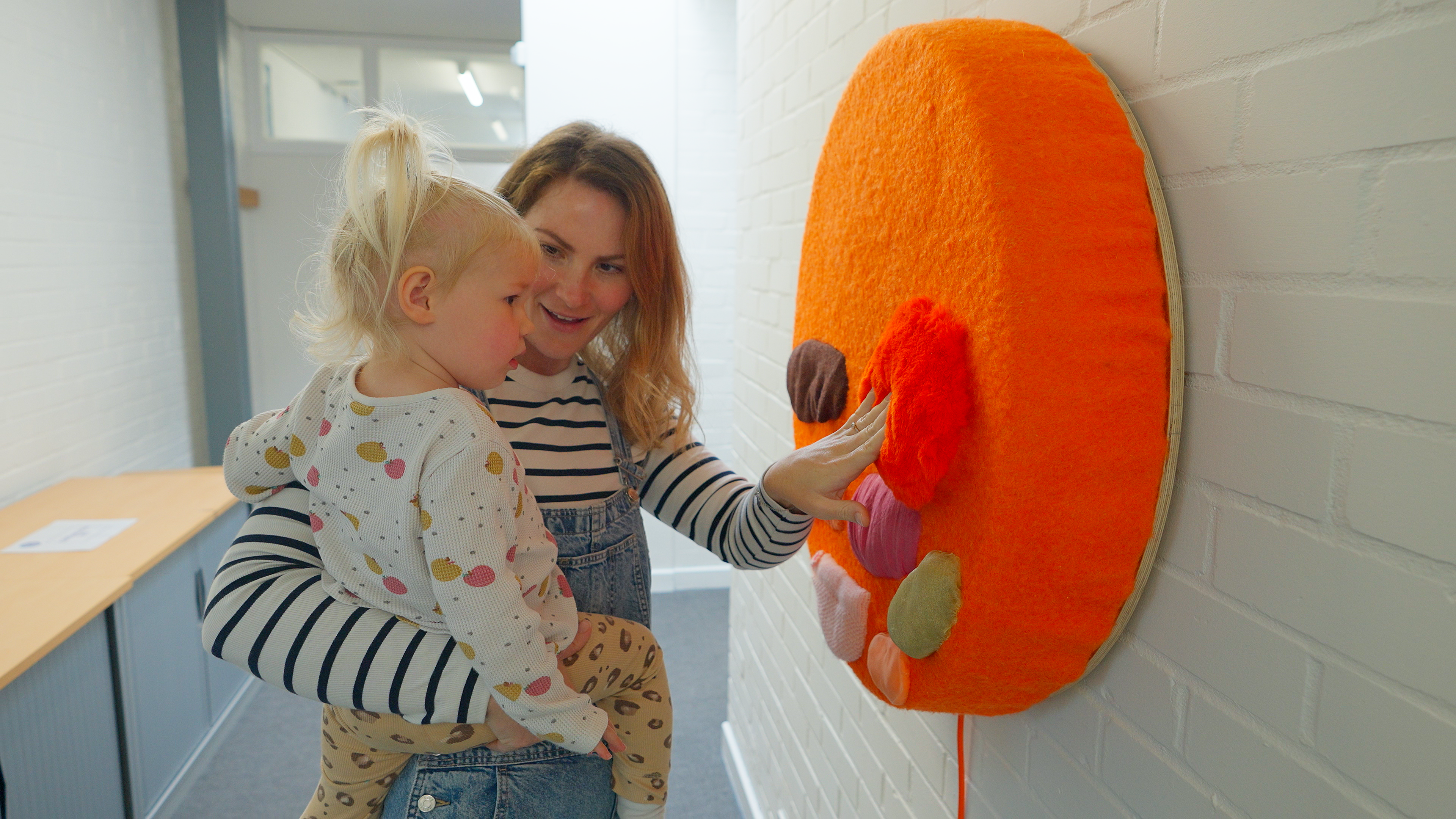 A mother and infant engaging with an installation. The mother is touching an orange circle with different colour shapes and textures on it.