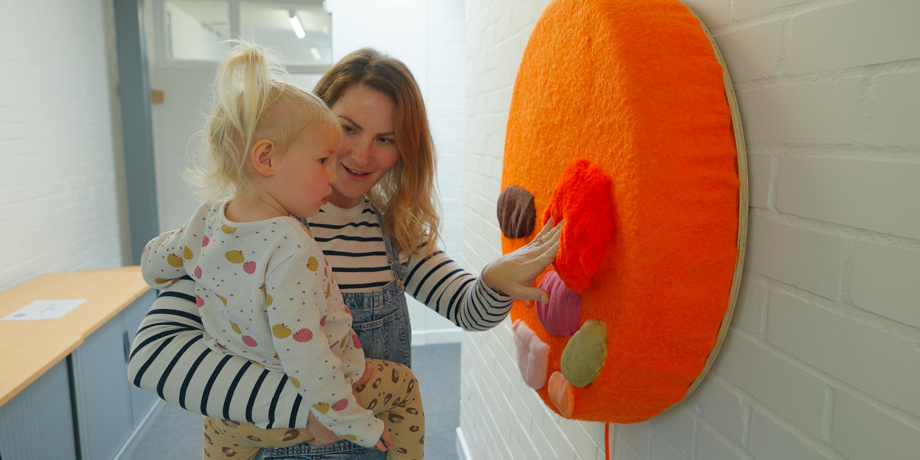 A mother and infant engaging with an installation. The mother is touching an orange circle with different colour shapes and textures on it.